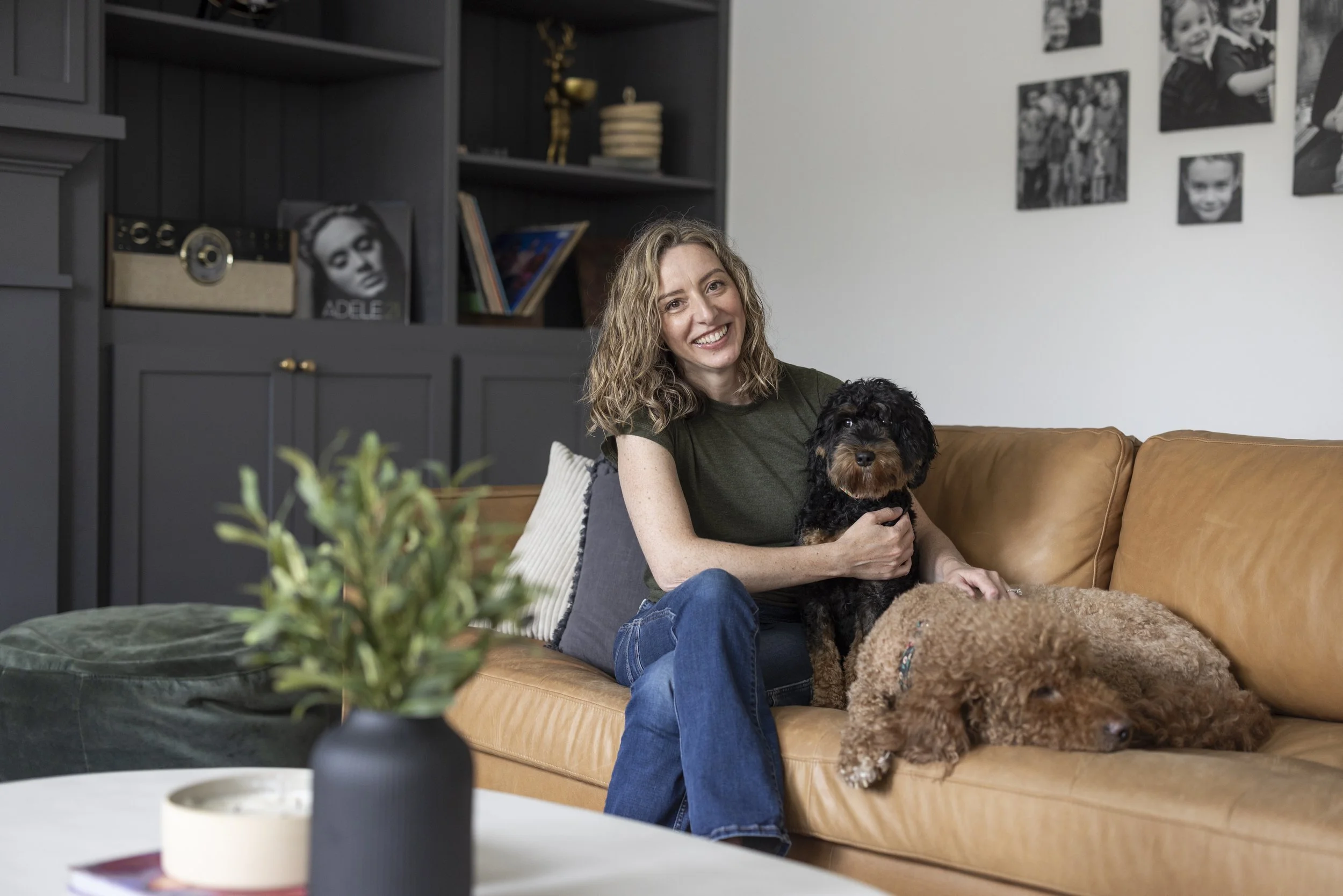 Tricia Caldwell and her labradoodle on a tan leather sofa in a finished living room with dark built-in shelving.