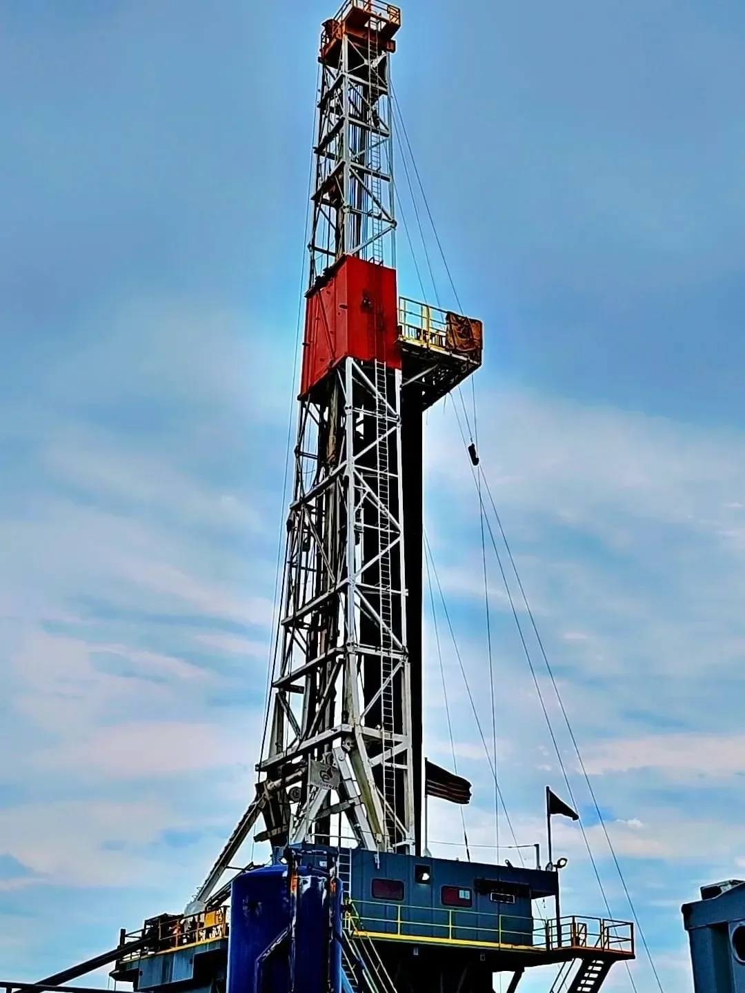 An oil drilling rig with a tall metal tower, orange and black components, and a platform at the bottom, set against a partly cloudy blue sky.