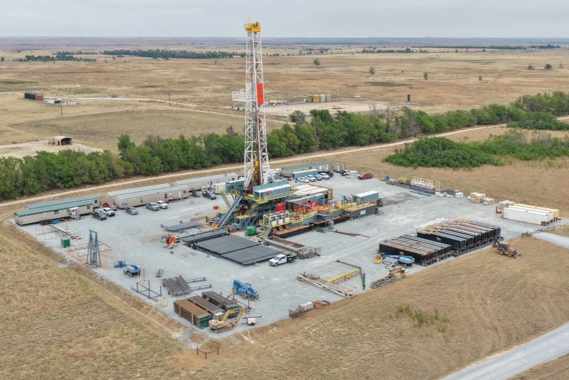 An oil drilling rig surrounded by trees and open fields in a rural area.