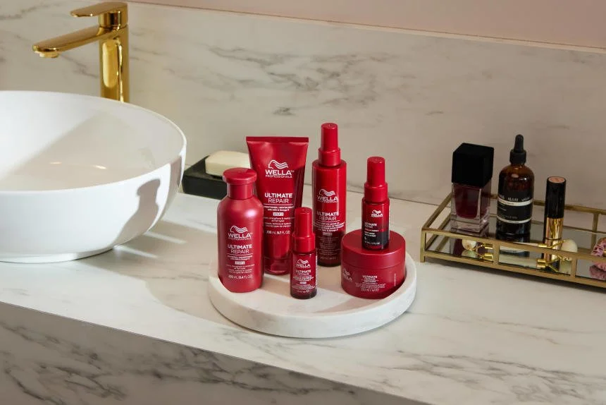 Verschiedene Wella Haarpflegeprodukte auf einer weißen Kuchenplatte auf einem Waschtisch mit goldener Armatur im Badezimmer, daneben eine goldene Kosmetikablage mit weiteren Kosmetikprodukten.