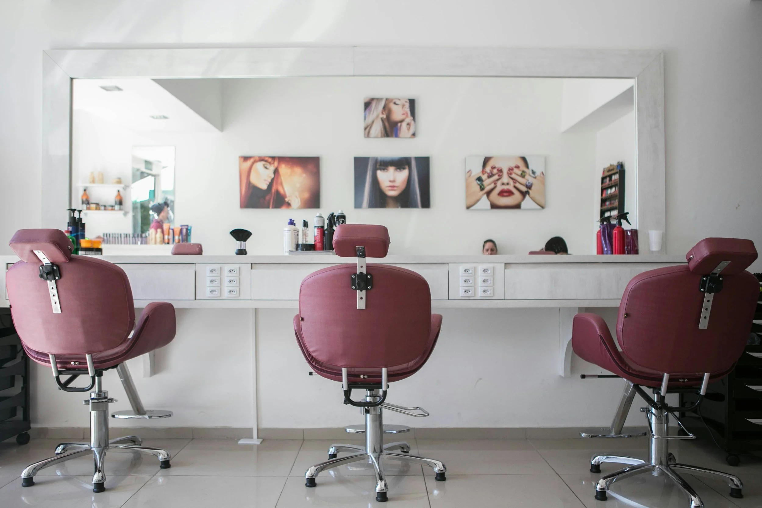 Ein Friseursalon mit einem großen Spiegel, Pastellrosa Friseurstühlen, einem weißen Waschbecken, verschiedenen Stylingprodukten auf einem Regal und Wandbildern von Models mit unterschiedlichen Frisuren und Make-up.