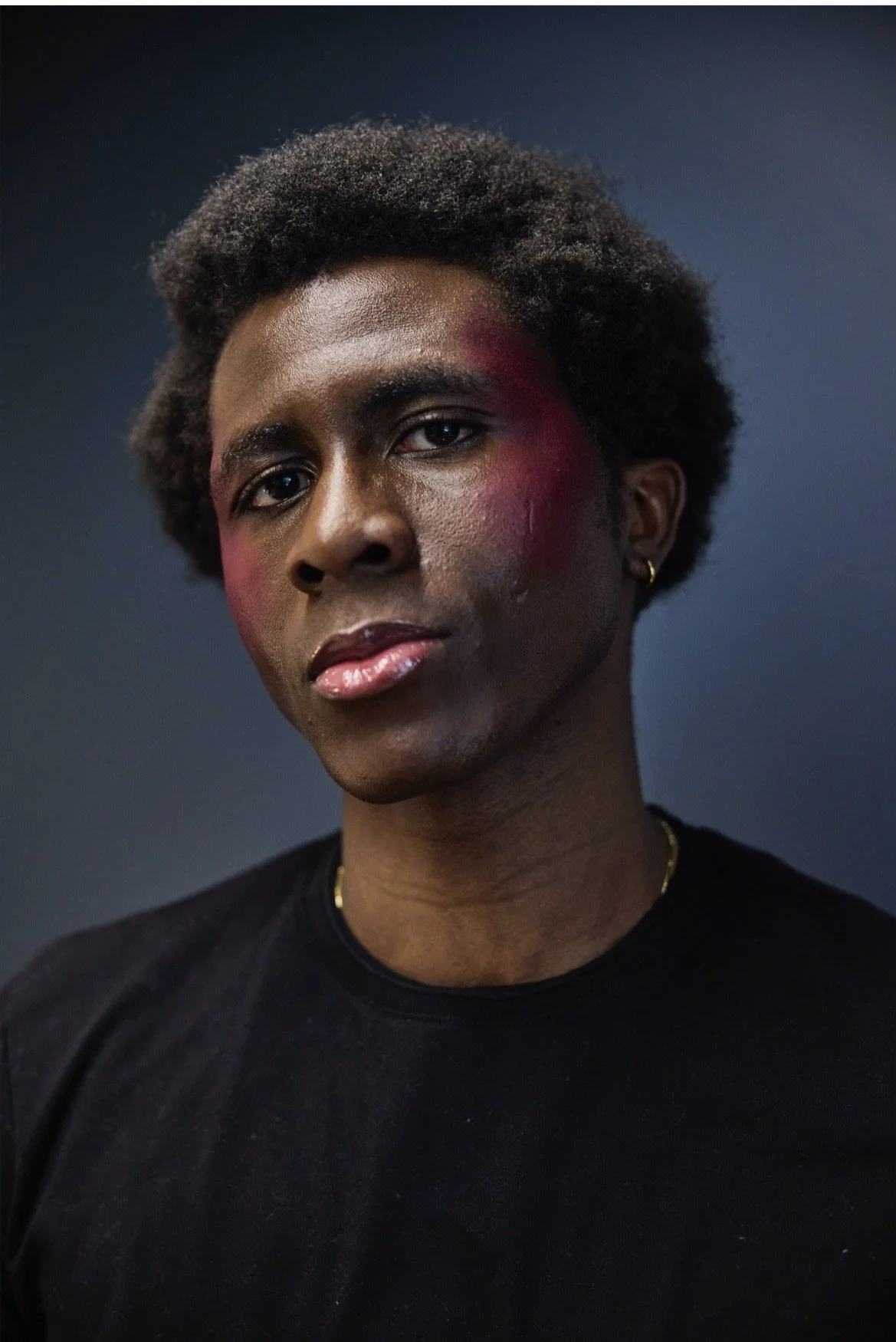 Porträt einer schwarzen Frau mit kurzer, lockiger Frisur, ernstem Gesichtsausdruck, makeup mit rotem Rouge auf den Wangen, schwarze Kleidung, einfache goldene Ohrringe und Kette, dunkler, unscharfer Hintergrund.