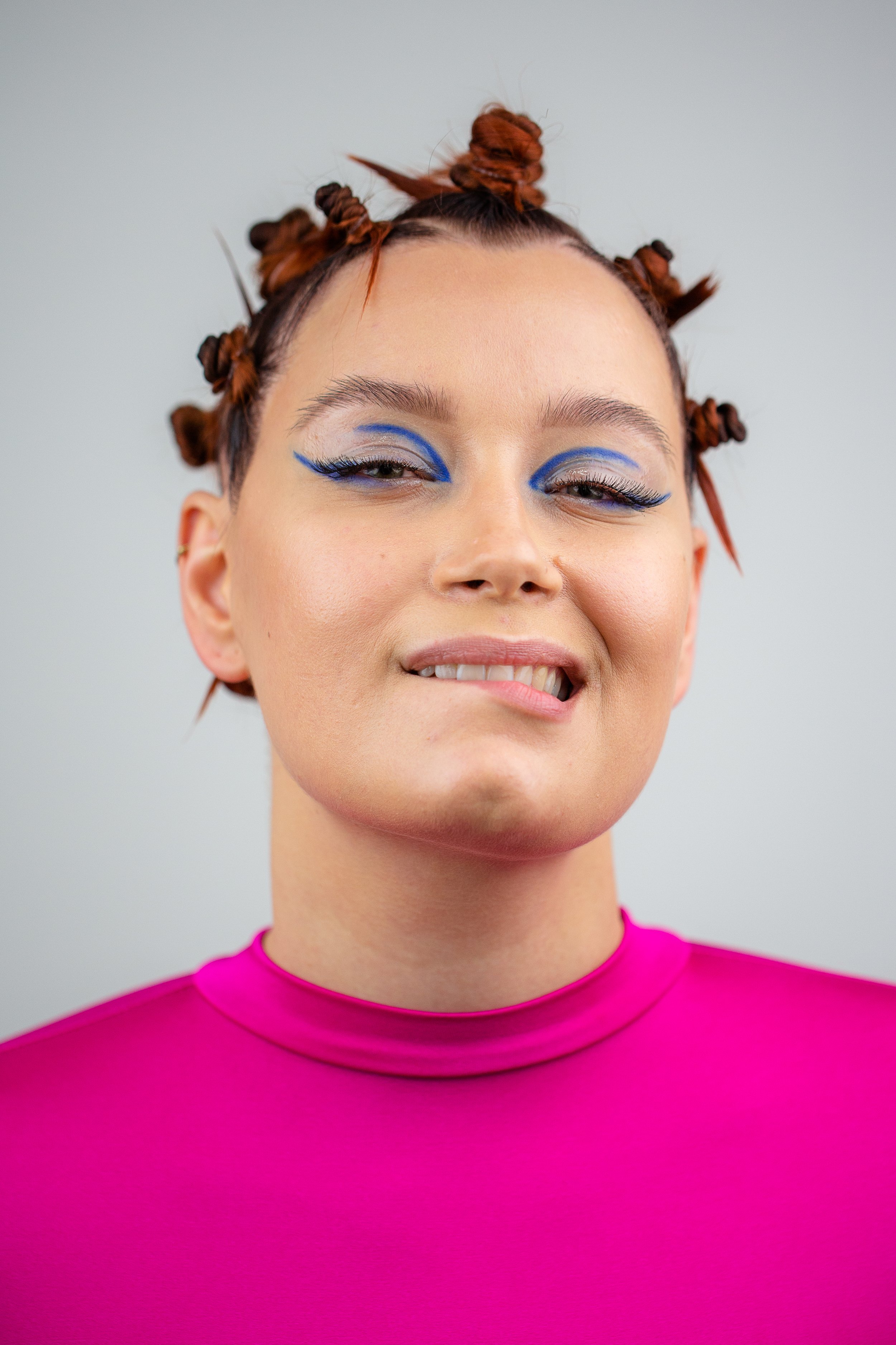 Junge Frau mit künstlerischem blauen Augen-Make-up, kurzen roten Locken im Zopf, trägt ein pinkes Oberteil, lächelt leicht und blickt teilweise in die Kamera.