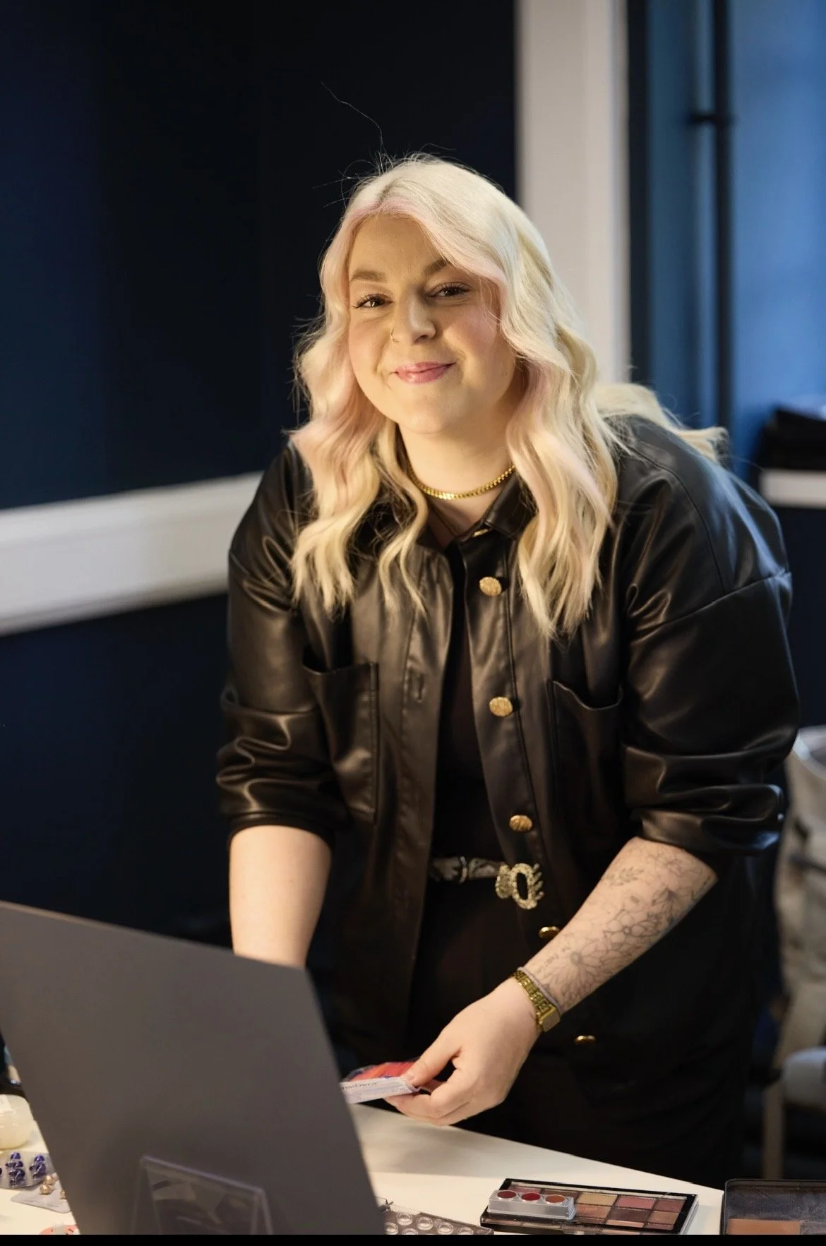 Eine junge Frau mit blonden, gewellten Haaren, in dunkler Kleidung, die vor einem Tisch mit Make-up-Produkten sitzt und lächelt.