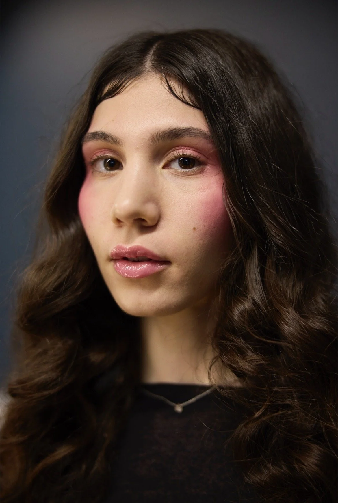 Nahaufnahme eines jungen Mädchens mit braunen, welligen Haaren, smoky Augen-Make-up und glanzlosem Lippenvolumen.