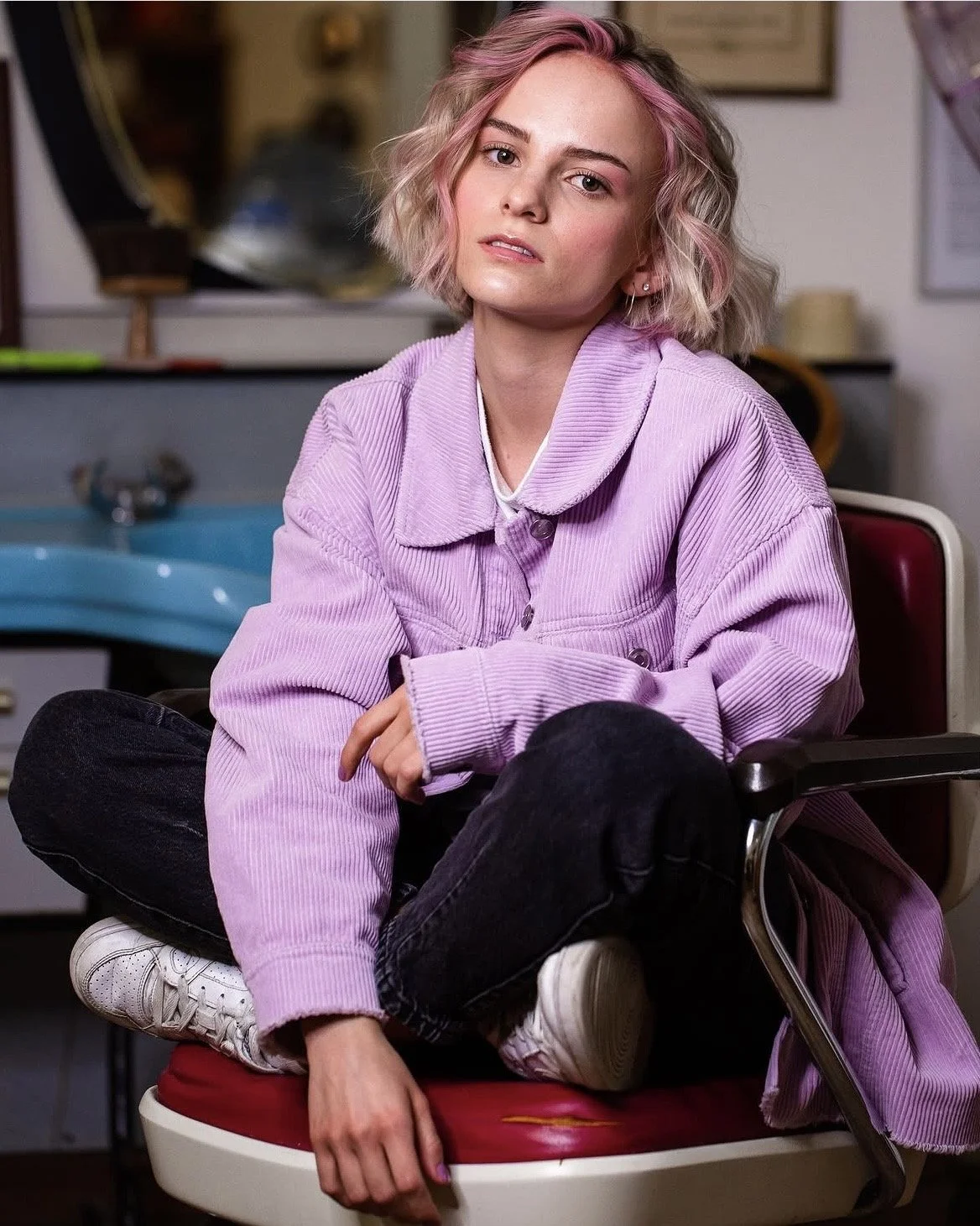 Junge Frau mit pinkblondem, lockigem Haar, in lila Jacke, sitzt in einem Rollstuhl in einer Einrichtung, im Hintergrund Küche und Therapieraum.