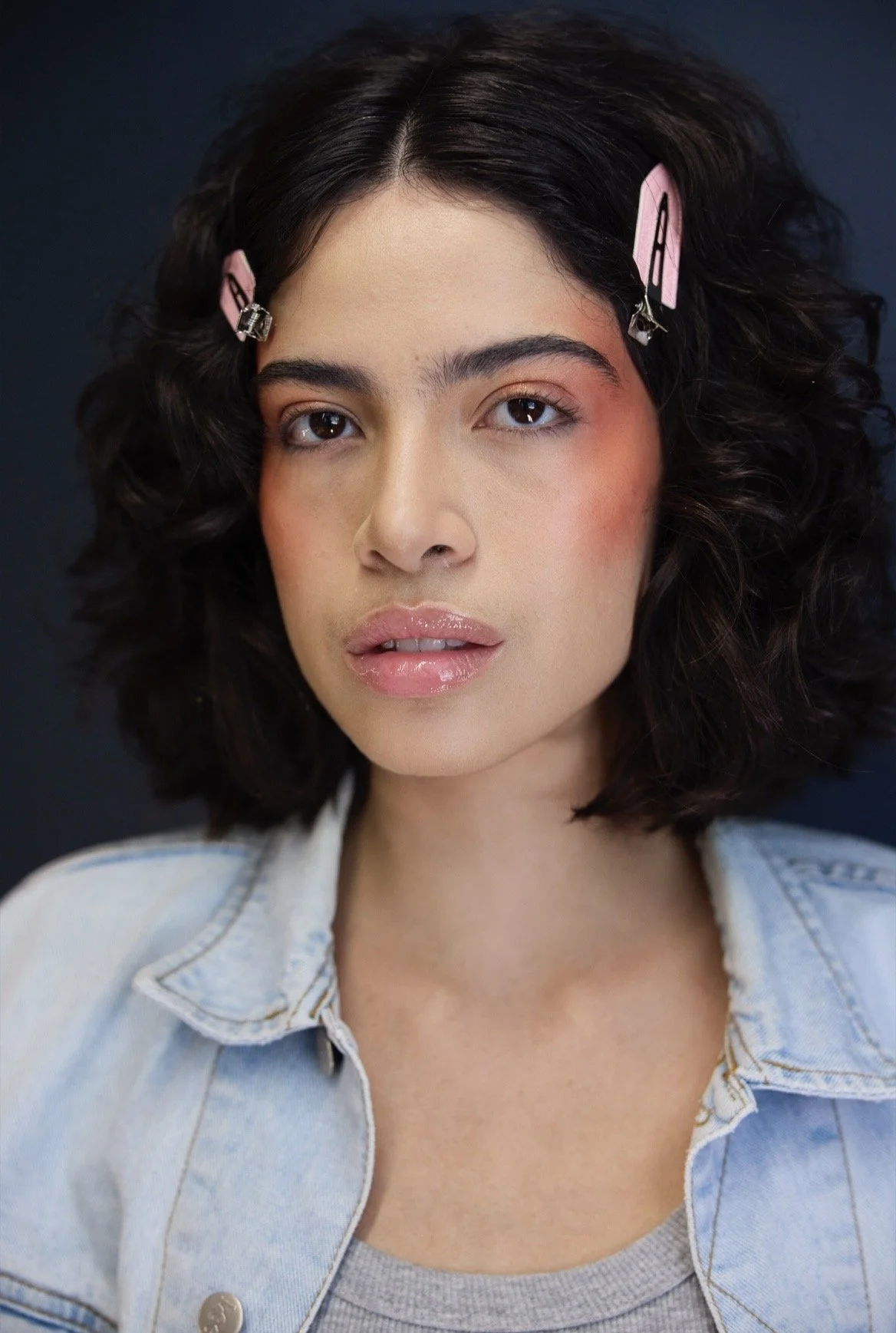 Nahaufnahme einer jungen Frau mit lockigem schwarzen Haar, pinken Haarspangen, heller Haut, natürlichem Make-up, Jeansjacke und grauem Top vor dunklem Hintergrund.