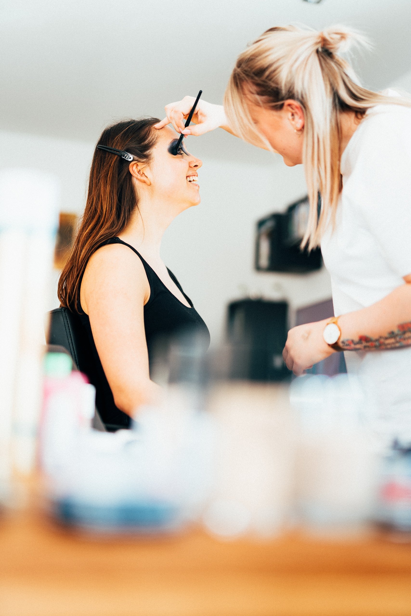 Eine Frau wird von einer Stylistin für ein Make-up geschminkt, sie sitzt im Sessel und lächelt.