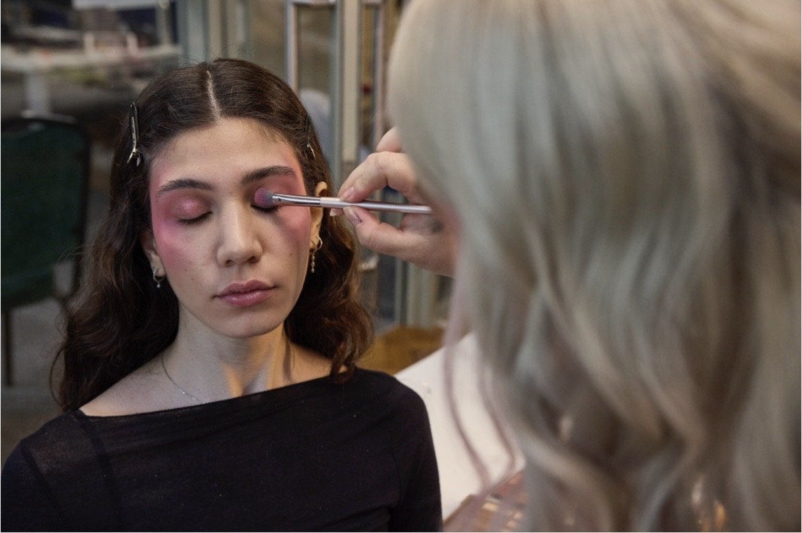 Eine Frau trägt Make-up auf die geschlossene Augenpartie einer jungen Frau auf, die mit dunklen Haaren und schwarzen Oberteil sitzt.