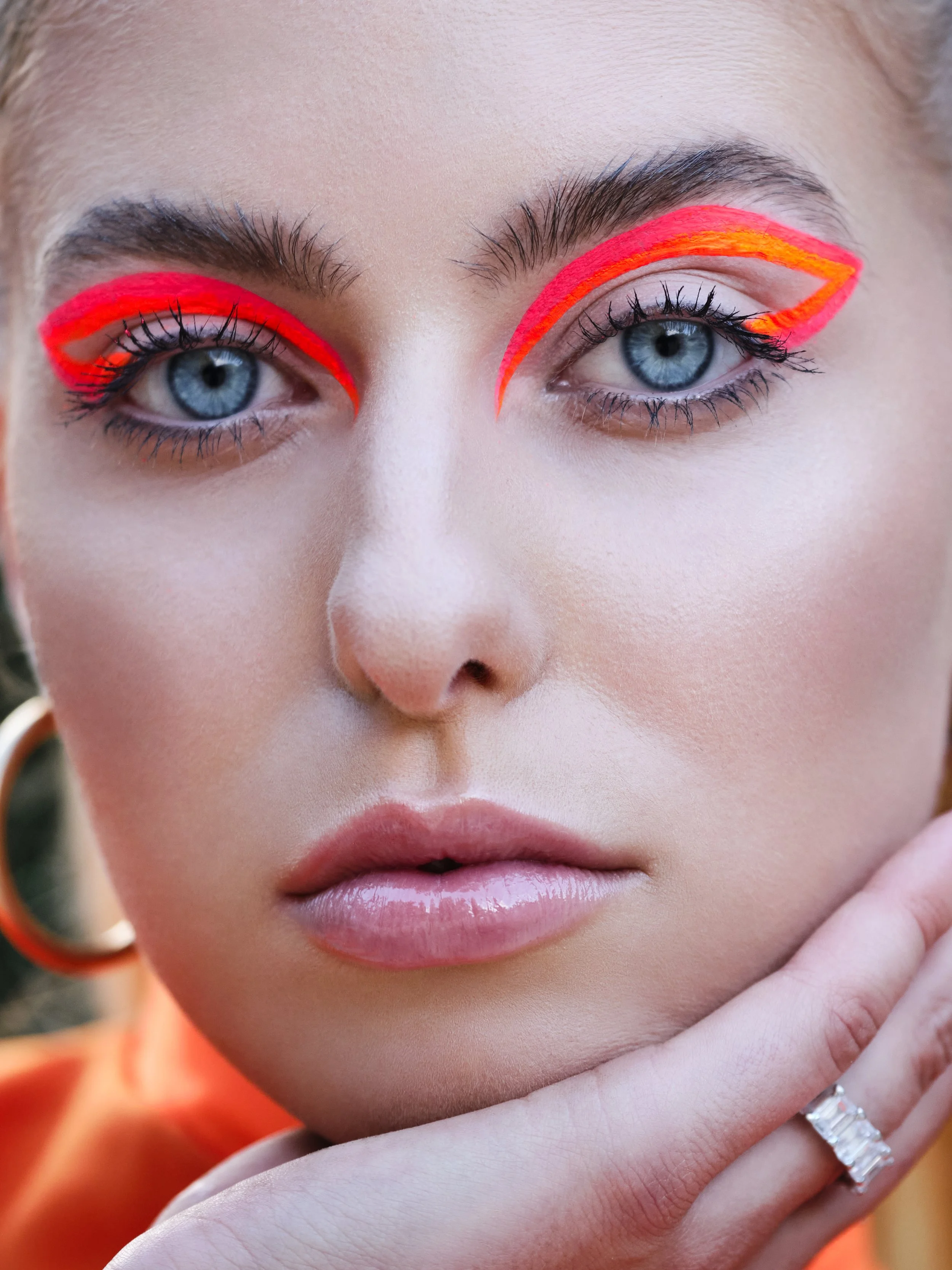 Nahaufnahme eines Gesichts mit blauen Augen, roten und orangefarbenen Eyeliner, Lippen mit hellrosa Lippenstift, Trägerin eines Ringes und einem orangefarbenen Kleid.