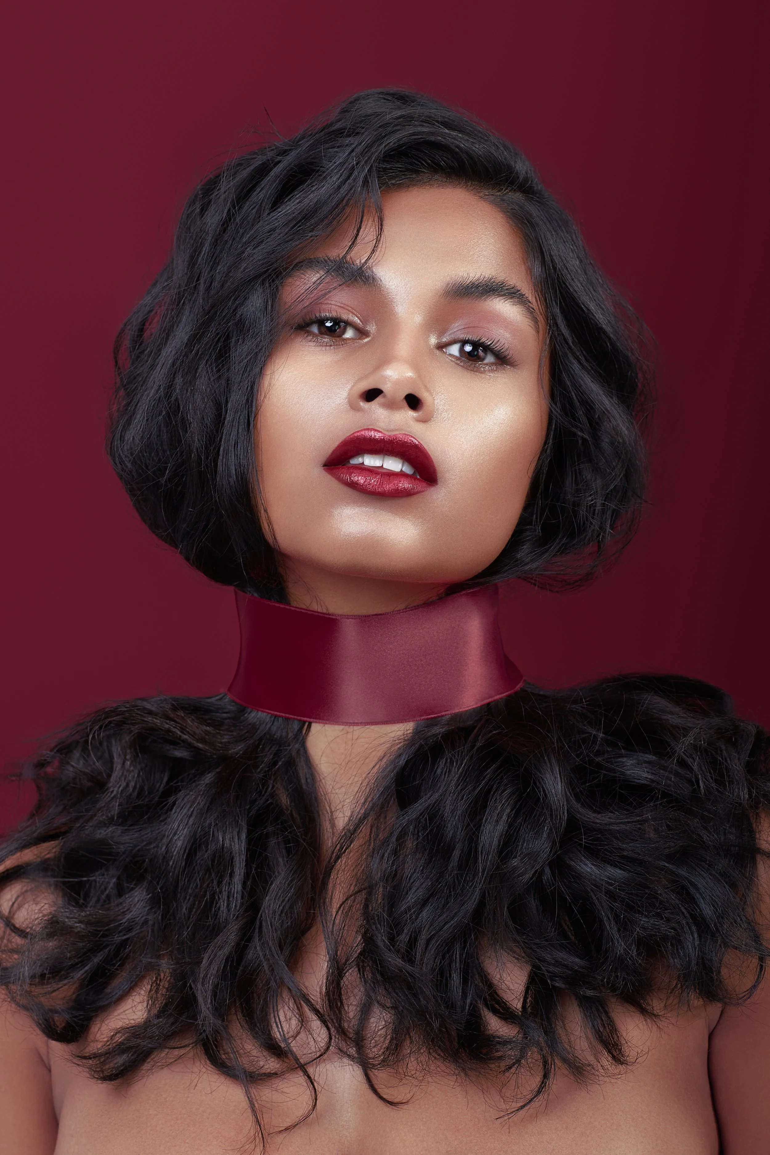 Nahaufnahme eines jungen Frauen mit dunklen, lockigen Haaren, rotem Lippenstift, dezenten Augen-Make-up, und einem maroon Lippenstift-Halsband vor einem dunkelroten Hintergrund.