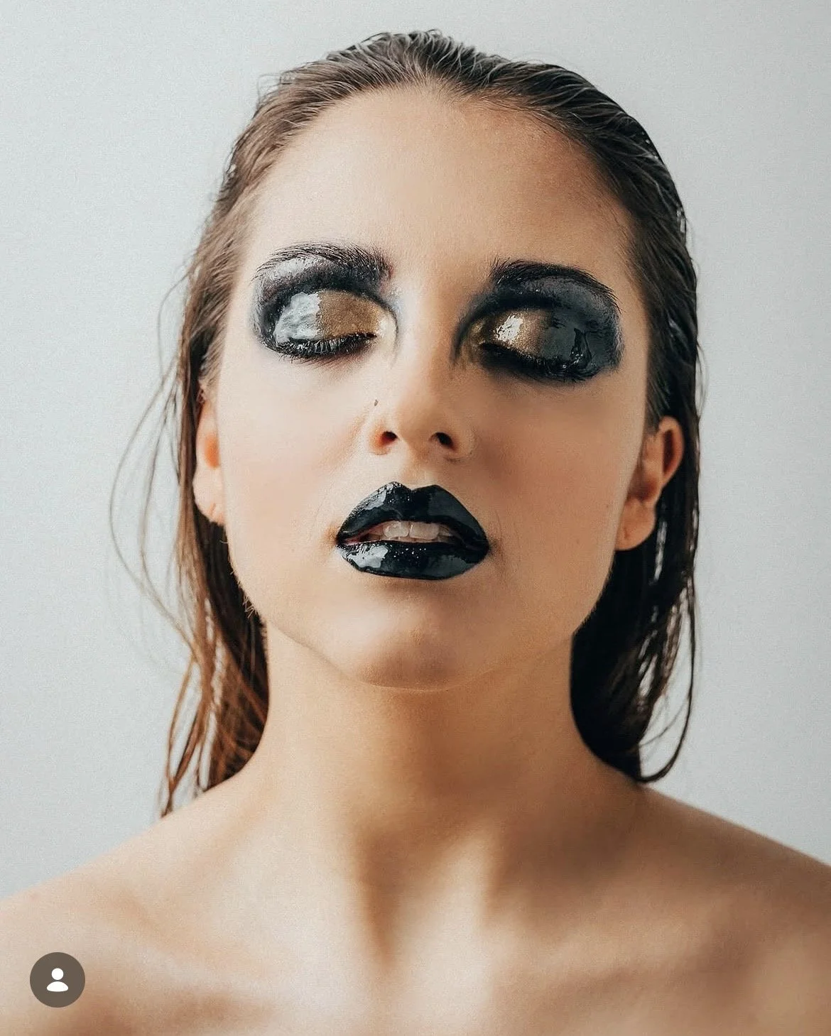 Junge Frau mit dramatischem schwarzen und goldenen Make-up, geschlossene Augen, leicht geöffneten Lippen, nackt Schulter, gegen weiße Wand blickend.