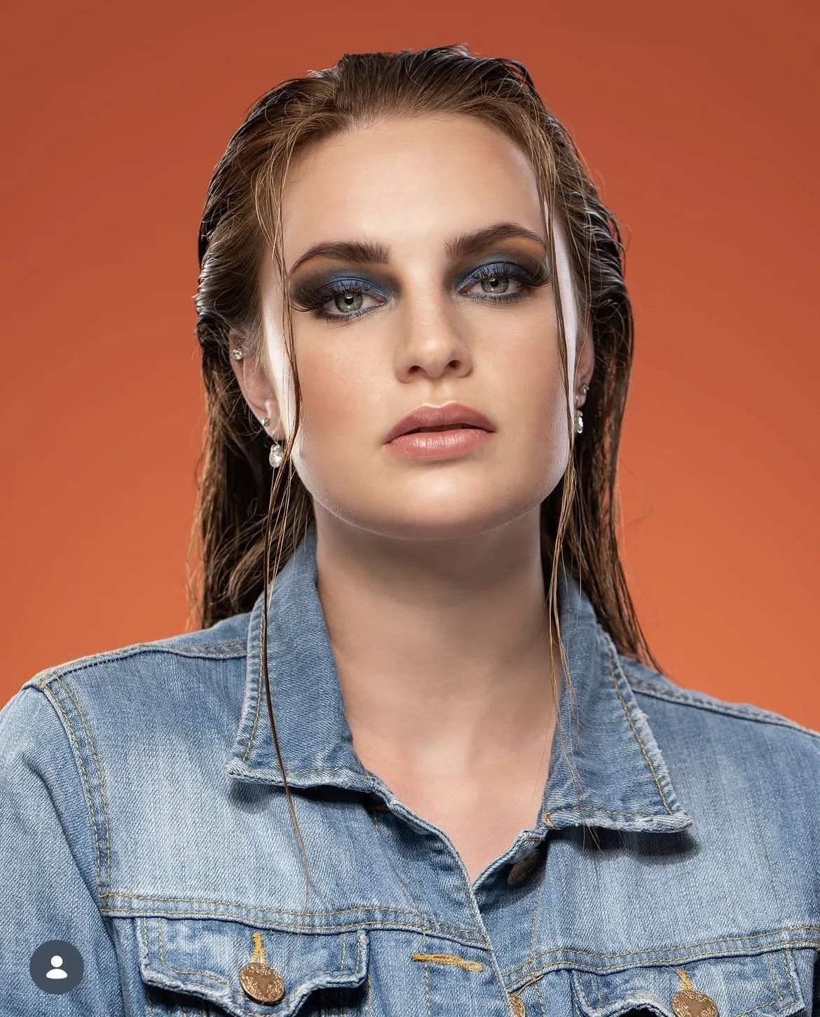 Nahaufnahme eines jungen mädchens mit blonden, nassen Haaren, trägt blaue Augen-Make-up, ein Jeanshemd und Ohrschmuck, vor orangefarbenem Hintergrund.