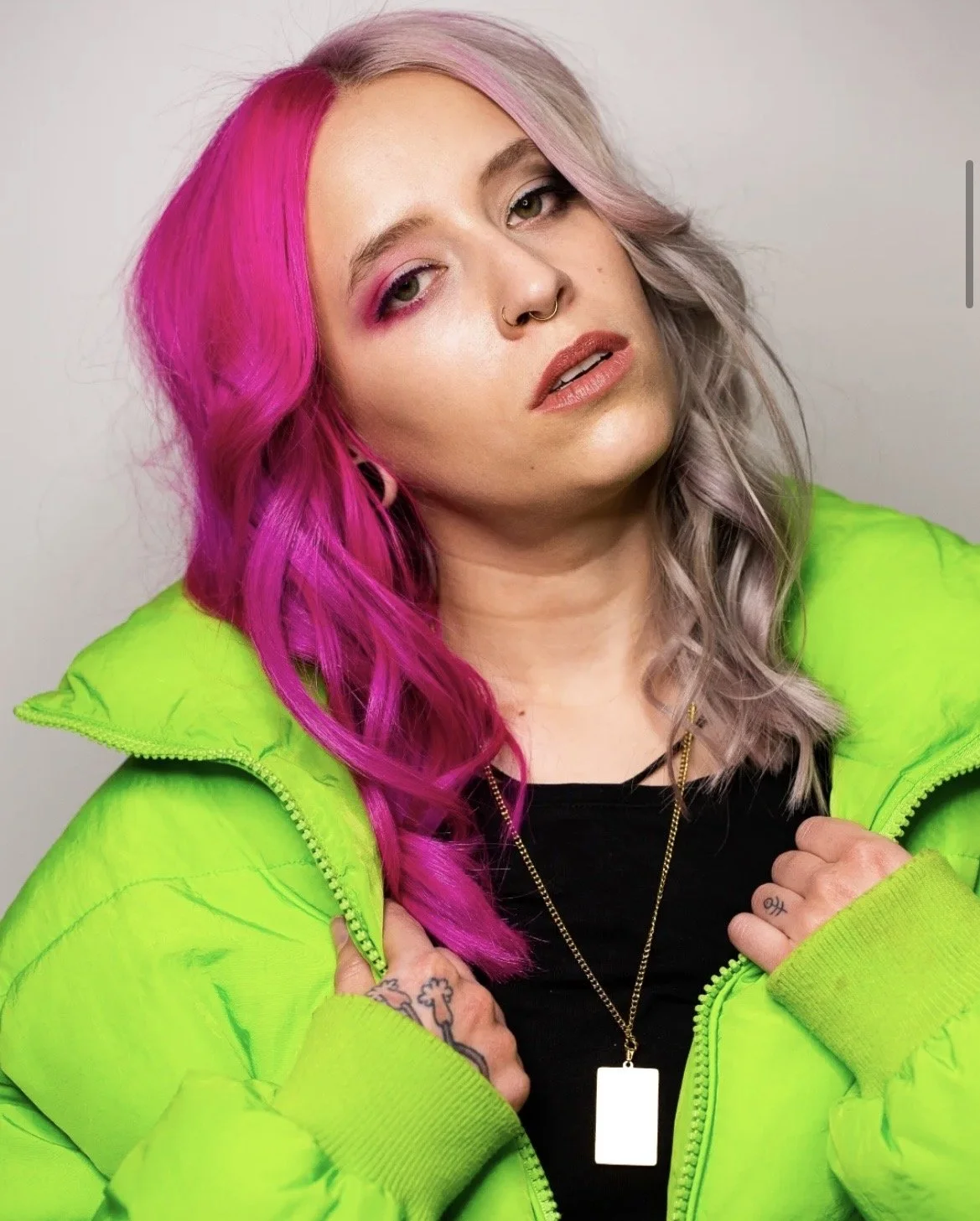 Porträt einer jungen Frau mit pinkem und lila Haar, die eine neon grüne Jacke trägt, vor neutralem Hintergrund.