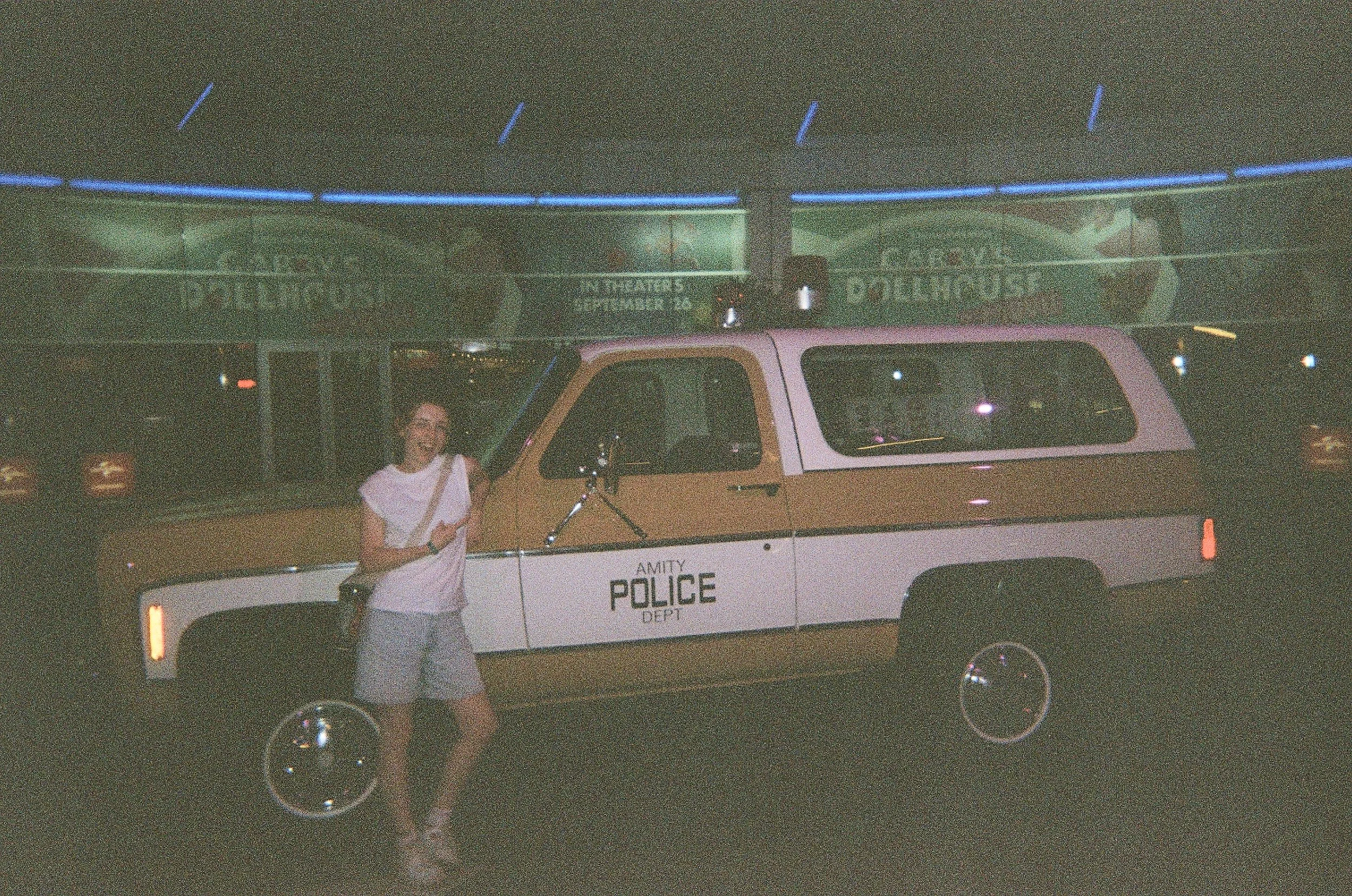A woman standing next to a police vehicle at night, with blue neon lights in the background.