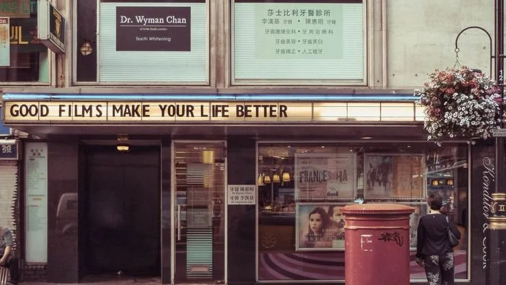 The storefront has a sign that reads, 'GOOD FILMS MAKE YOUR LIFE BETTER,' with a person standing in front of it and flowers hanging on the side.