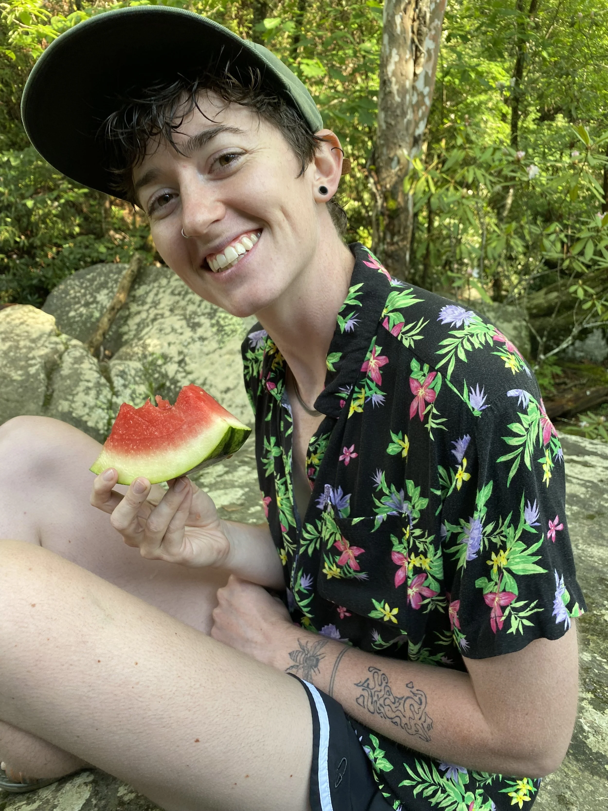 A smiling person with short dark hair and piercings, wearing a green cap and a black floral shirt, holding a slice of watermelon, and sitting outdoors in a wooded area with rocks and green foliage.