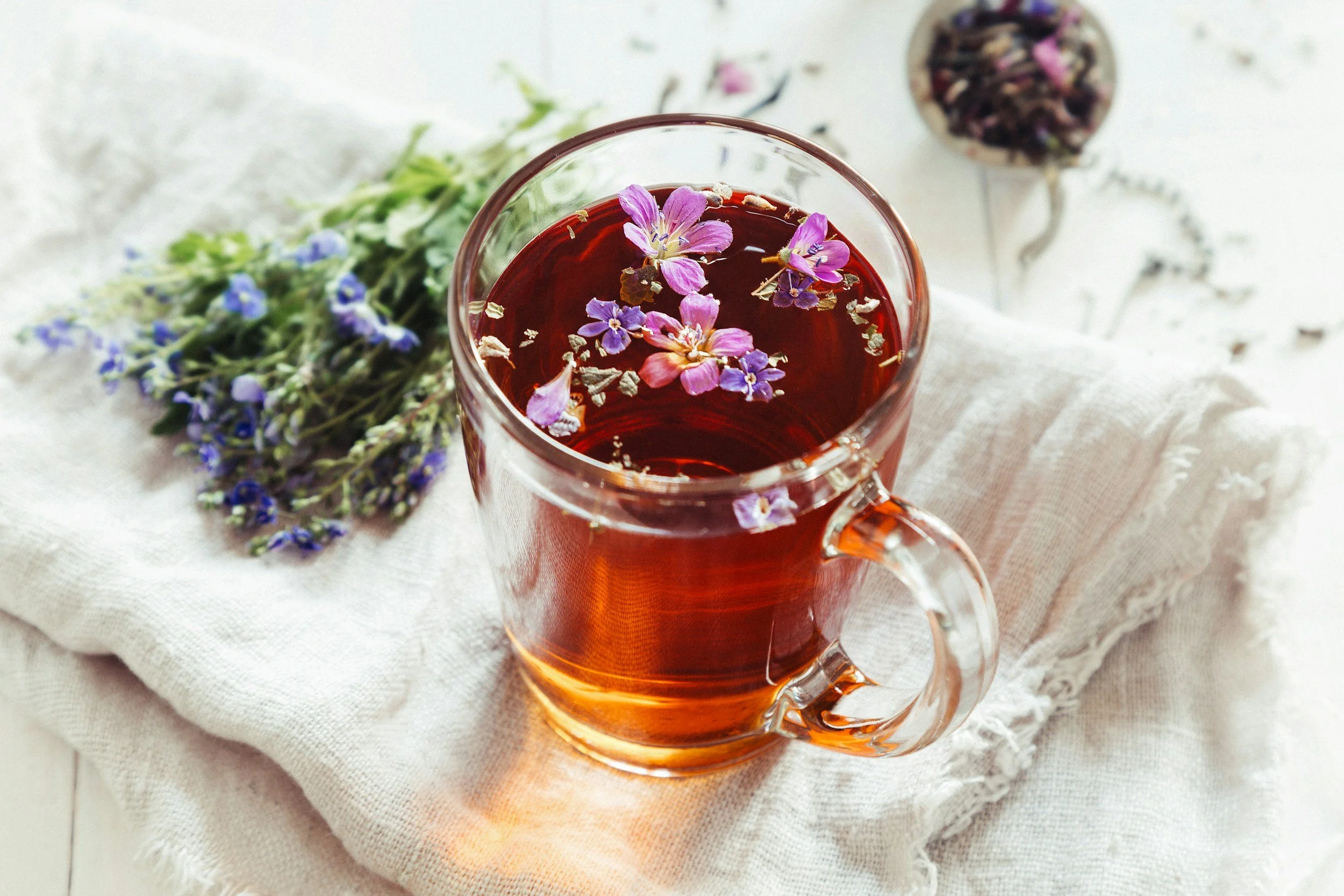 A clear glass mug filled with a reddish herbal tea, garnished with pink and purple flower petals floating on top. A bunch of purple flowers lying on a white textured cloth next to the mug, with a blurred container of dried flowers or herbs in the background.
