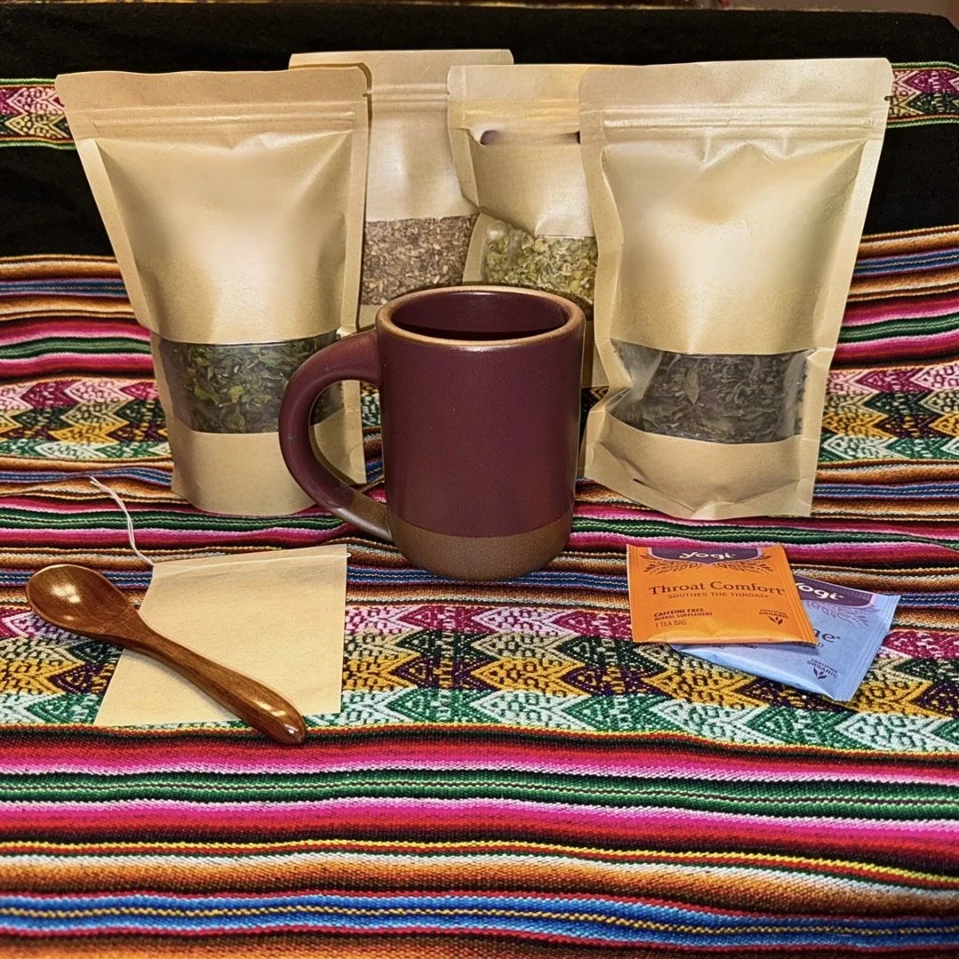 Three beige tea or herbal infusion pouches, a maroon and brown mug, a wooden spoon on a napkin, and various tea bags on a colorful woven textile.