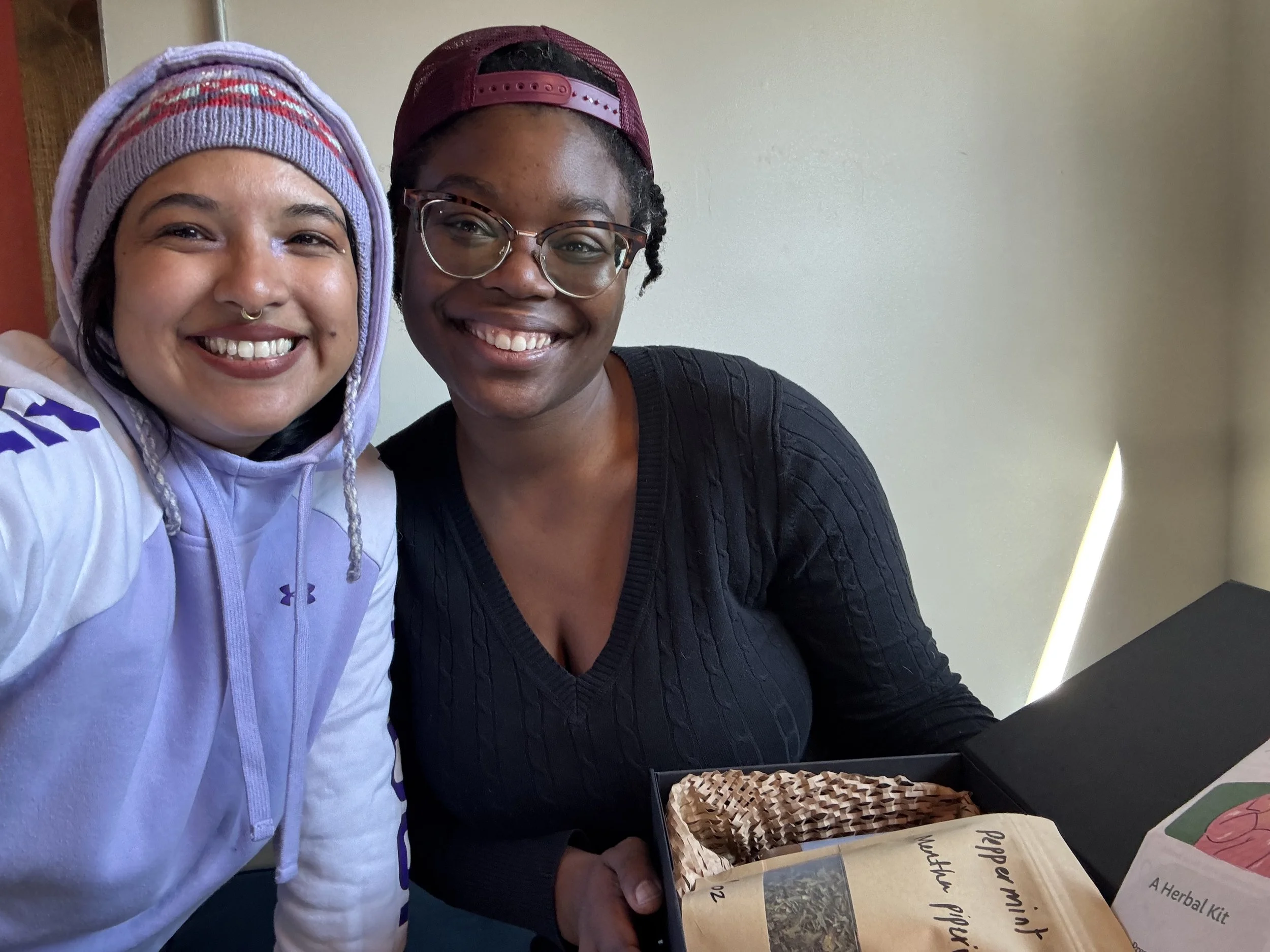 Two women smiling at the camera, one wearing a colorful beanie and the other wearing glasses and a maroon baseball cap, sitting at a table with herbal products and informational pamphlets in front of them.
