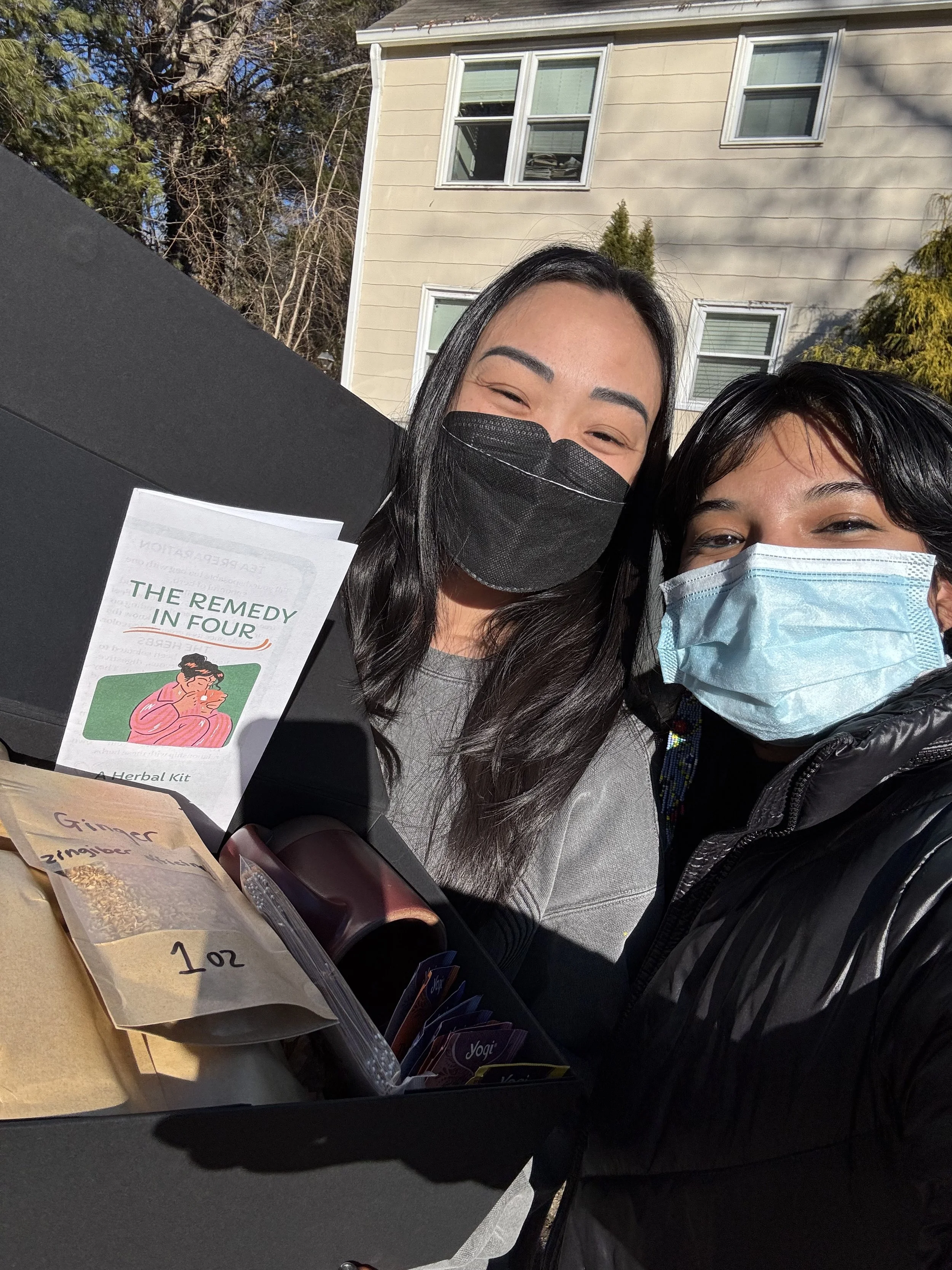 Two women wearing masks smiling behind a table with herbal and ginger products outdoors, with house and trees in the background.