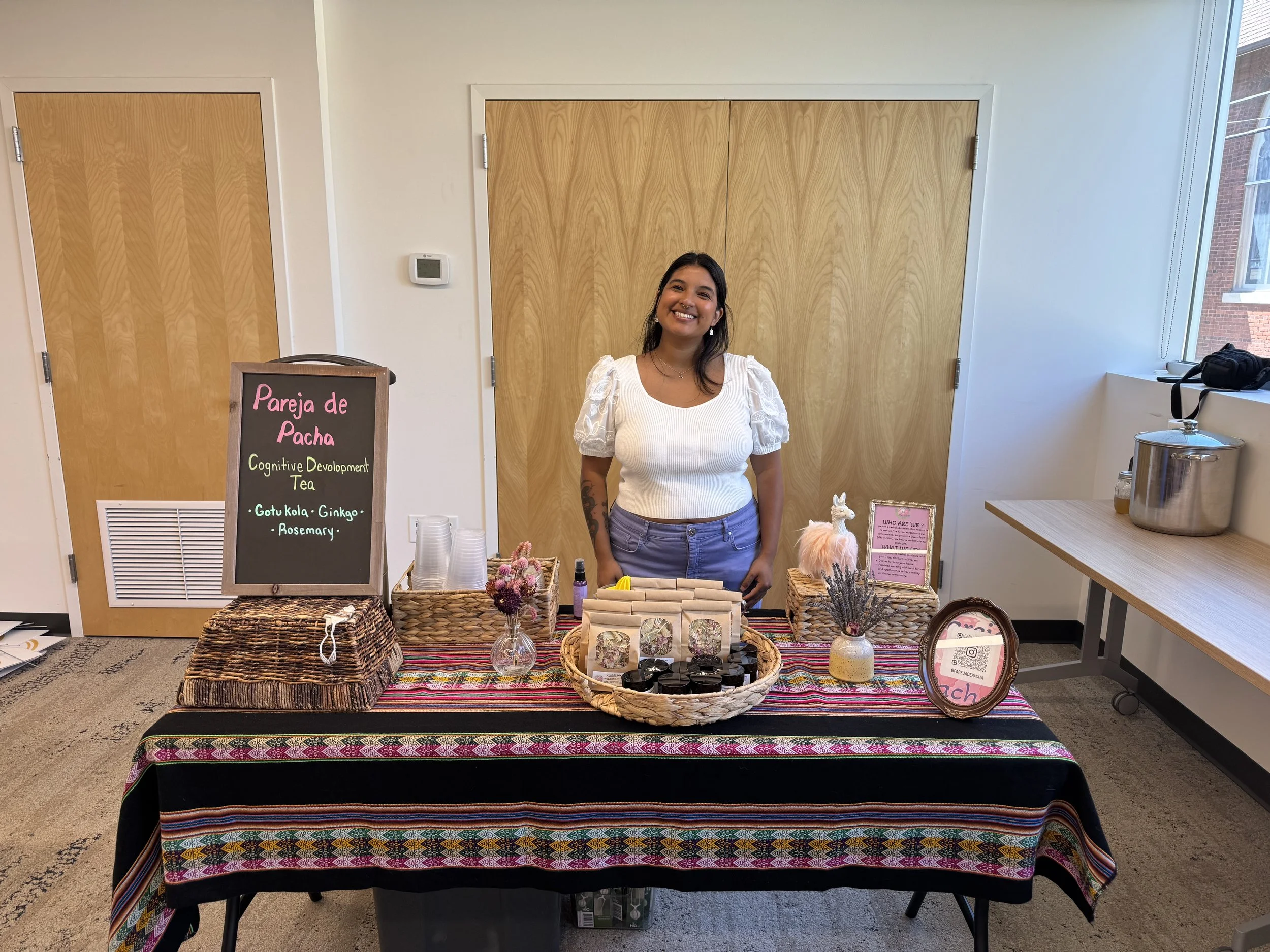 A woman standing behind a booth table displaying small products and decorative items, with a sign that reads 'Pareja de Pacha Cognitive Development Tea'.