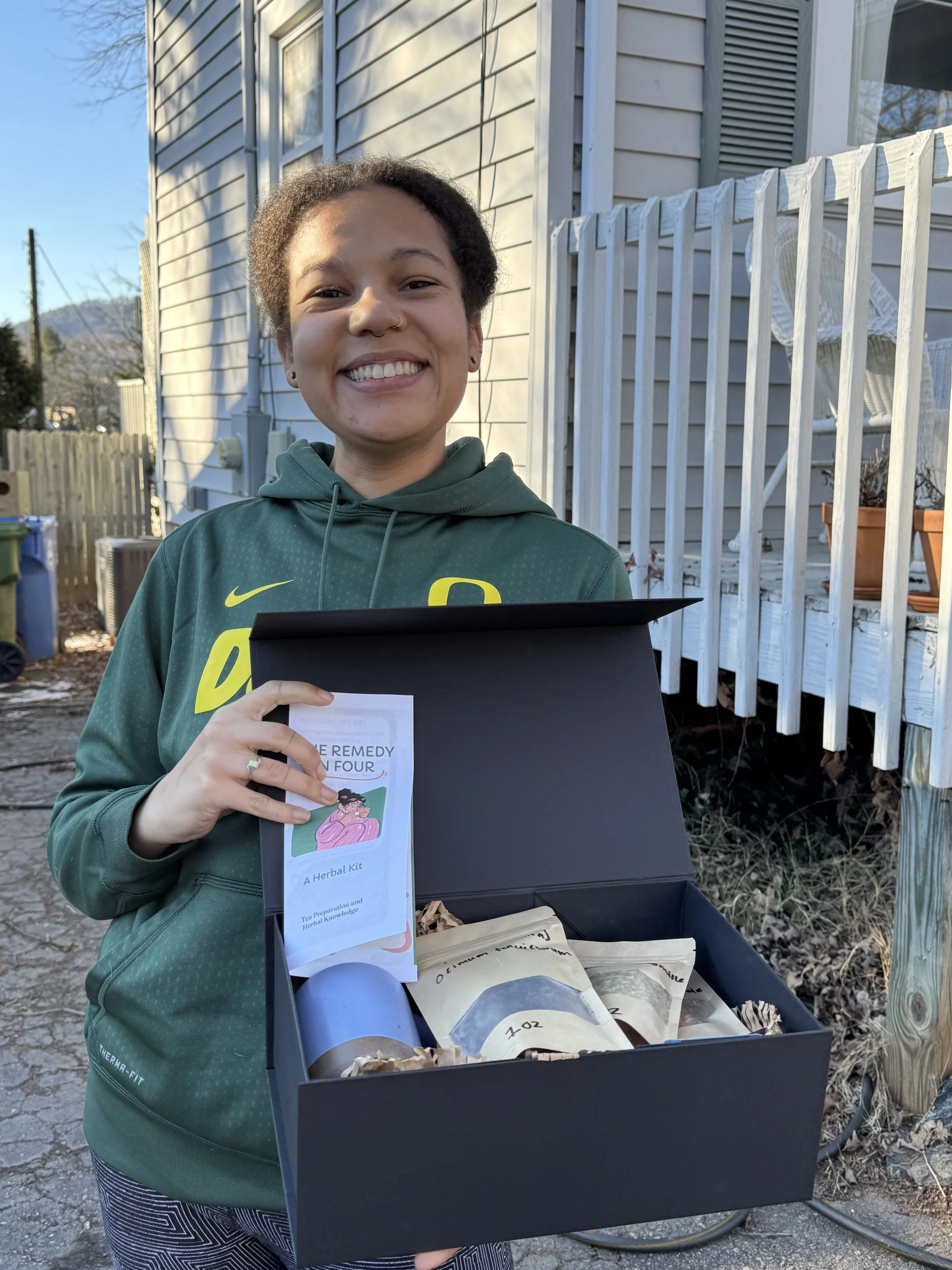 A young person smiling and holding a booklet in front of an open box with various papers and items. They are standing outside in front of a house with vinyl siding and a white wooden fence.