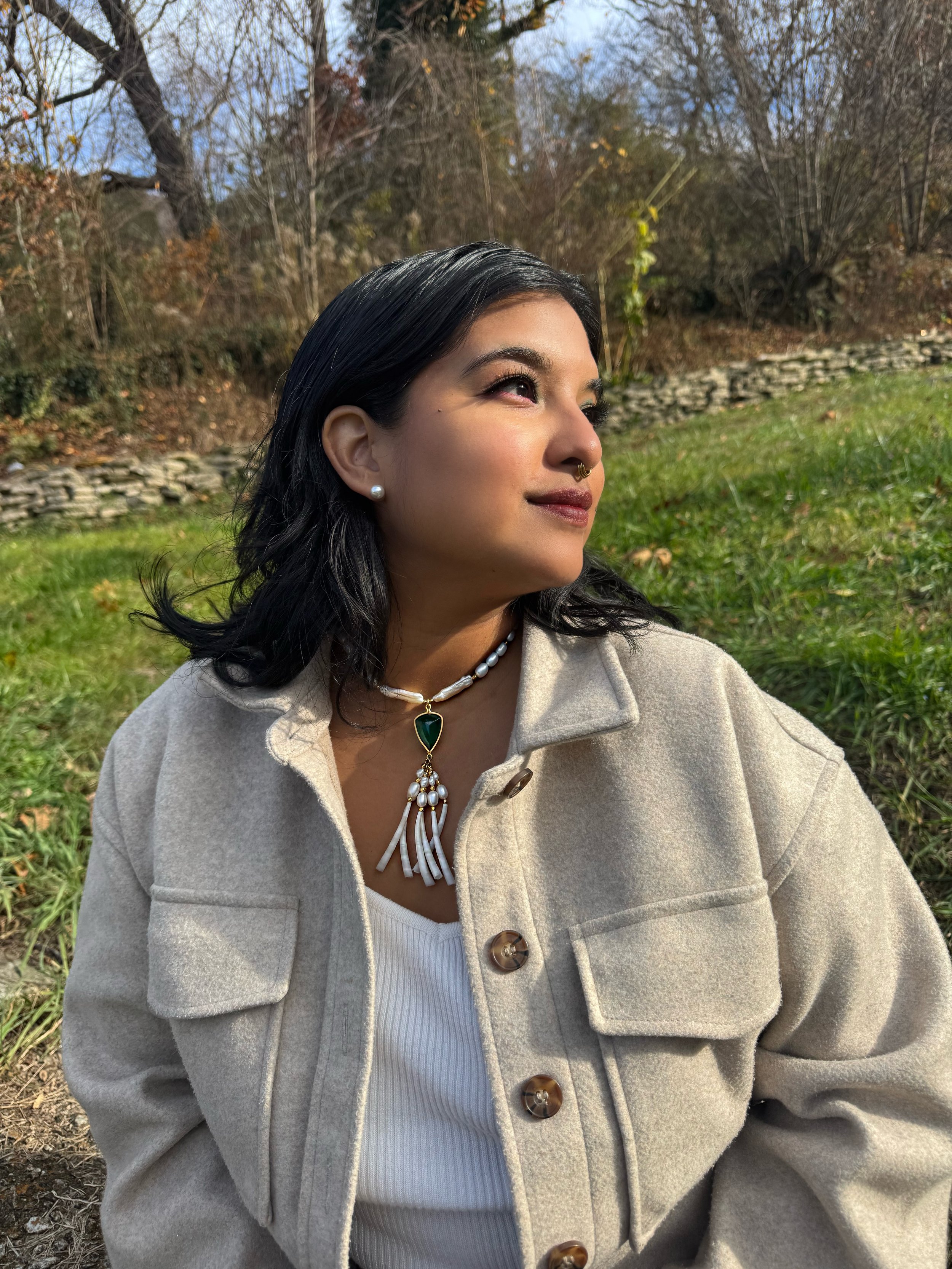 A woman with dark hair, wearing pearl earrings, a pearl necklace with a green pendant, and a beige coat, sitting outdoors with a backdrop of trees and grass on a sunny day.