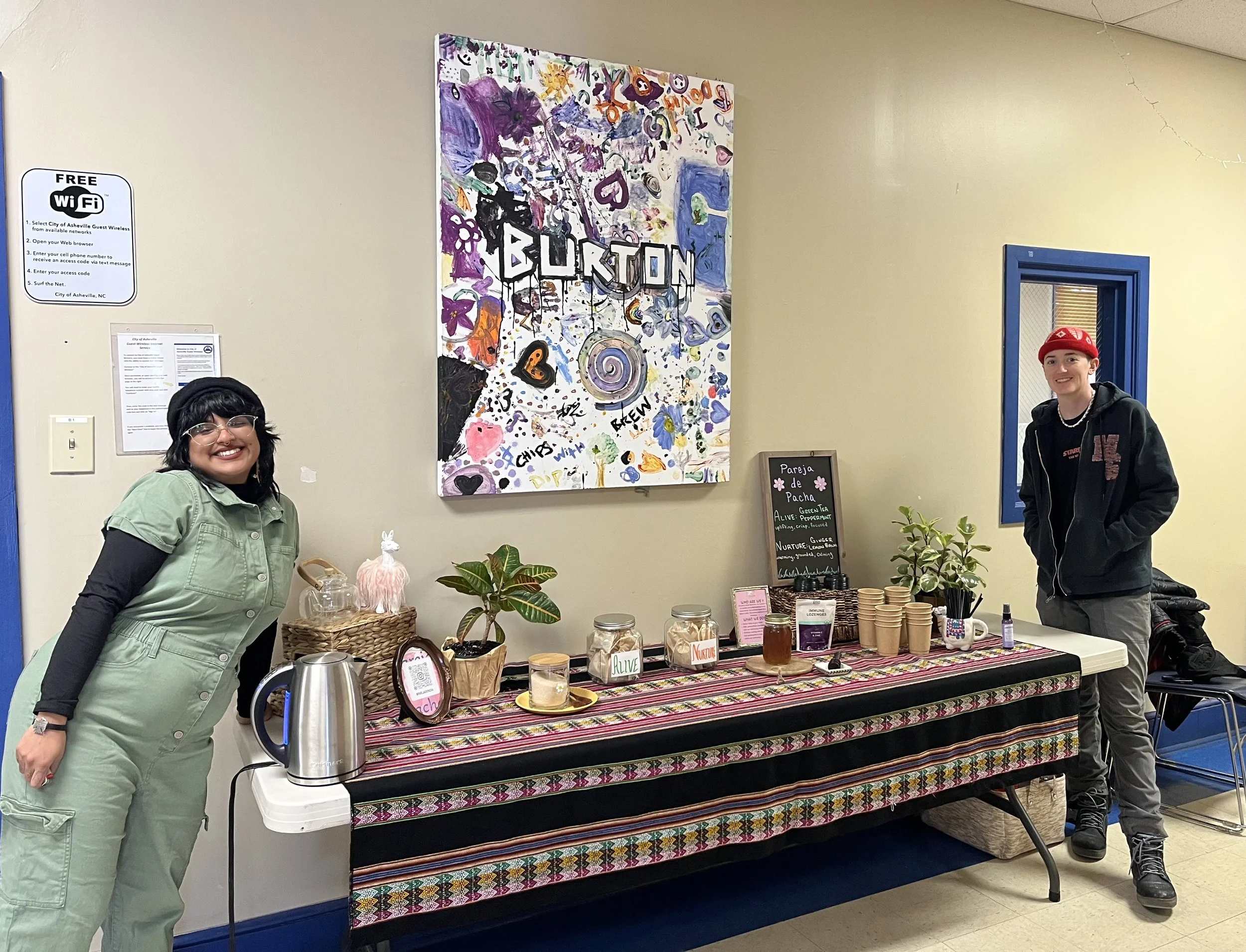 Two people smiling at a booth with herbal teas and wellness products, with a colorful abstract sign reading 'BURLTON' hanging on the wall behind them.