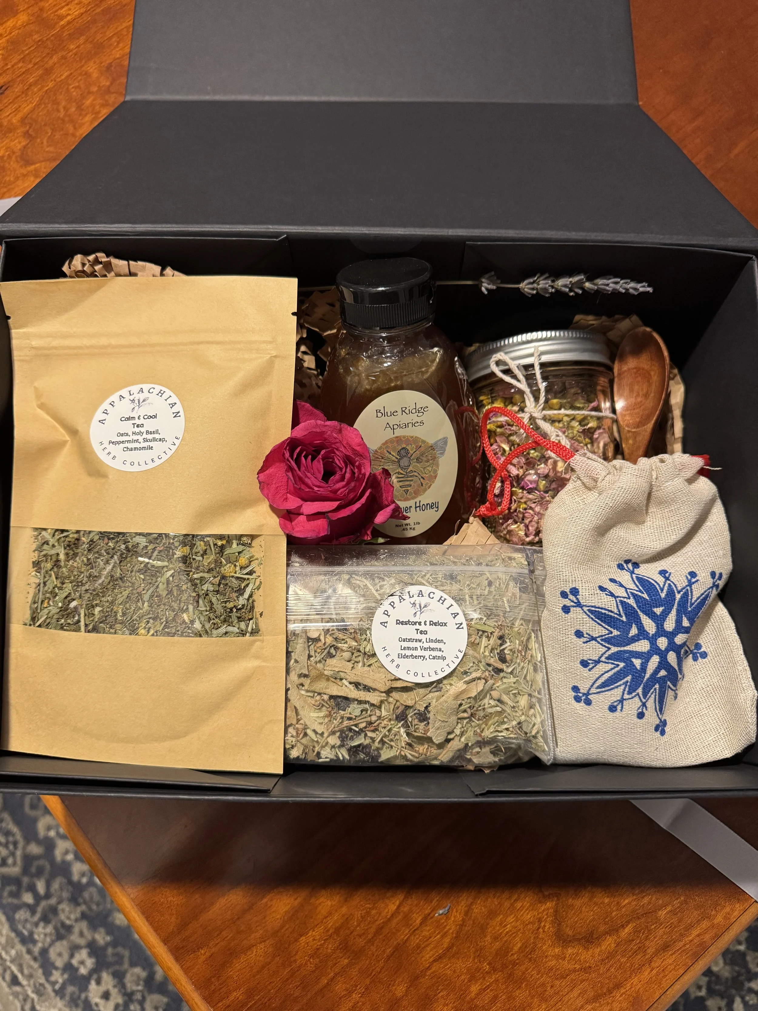A gift box with herbal tea, honey, dried herbs, a small bag, and a wooden spoon.