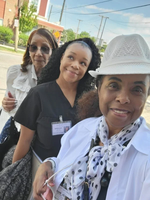 Three women standing outdoors, smiling for a selfie. One woman is wearing a white hat and polka dot scarf, another is in black scrubs with an ID badge, and the third woman is in a white blouse with sunglasses.