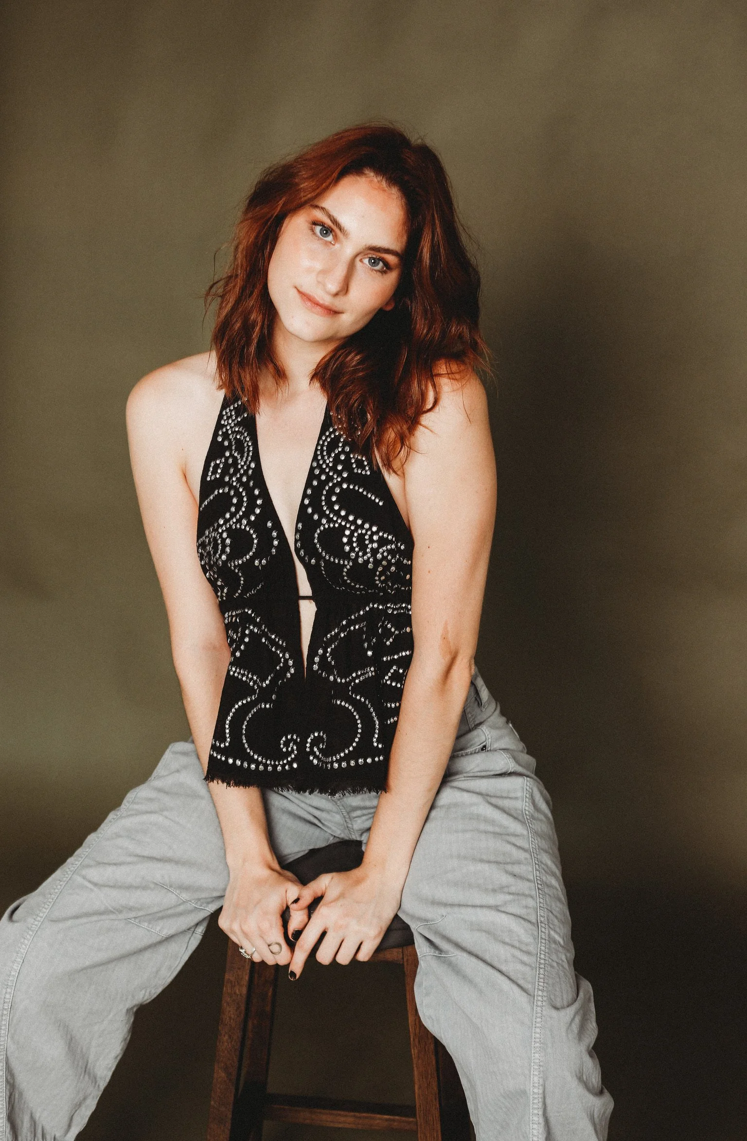 A young woman with shoulder-length wavy red hair and blue eyes, wearing a black sleeveless top with white embellishments, seated on a dark wooden stool against a plain dark background, looking at the camera with a slight smile.