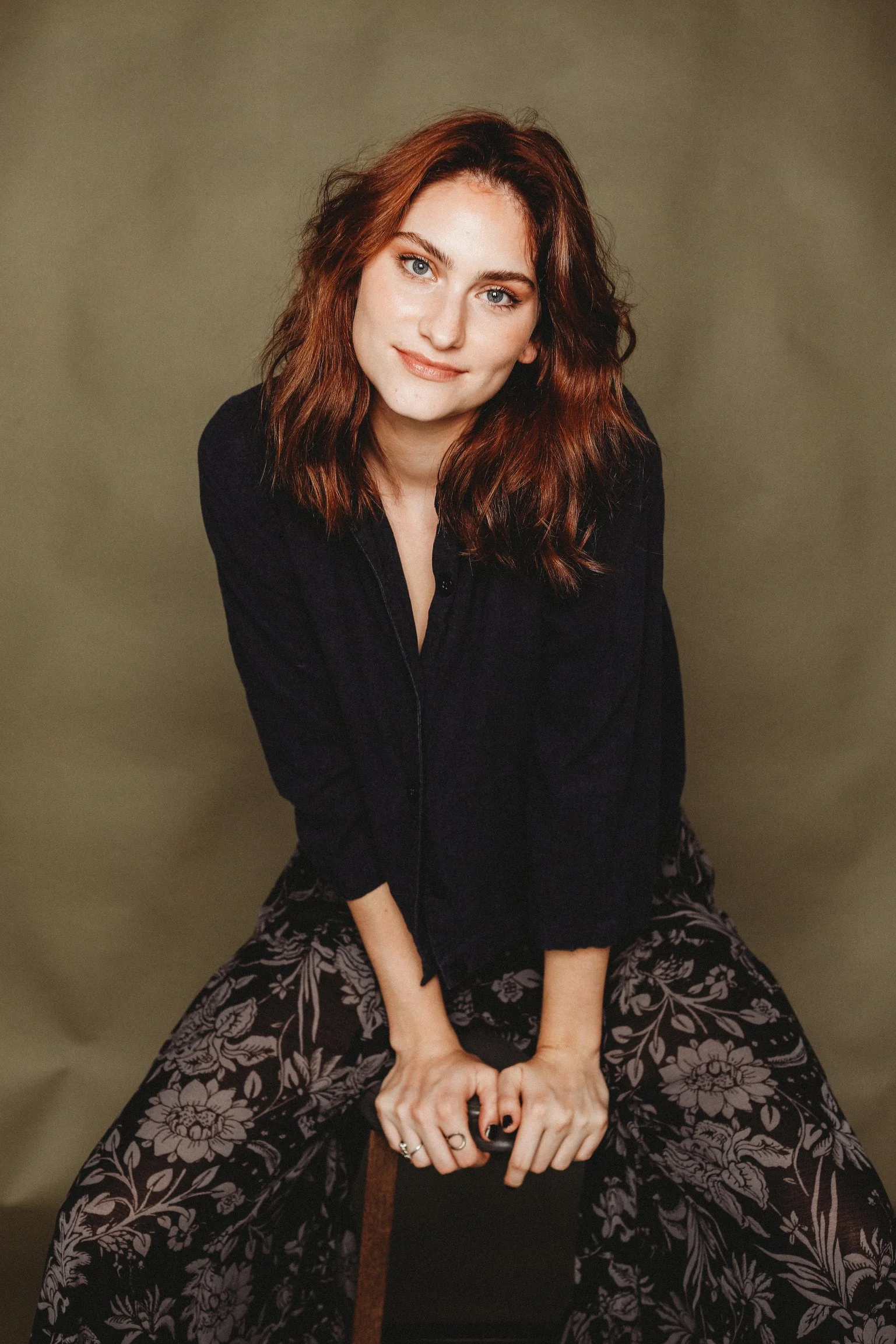 A woman with wavy red hair, blue eyes, and fair skin sitting on a chair against a plain background. She is wearing a black top and patterned wide-leg pants with floral design. She is looking at the camera with a gentle smile, leaning slightly forward with her hands resting on a round object.