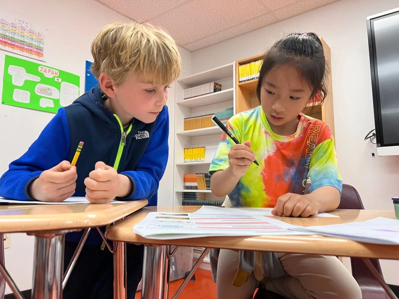 Two children, a boy with blonde hair wearing a blue jacket and a girl with dark hair wearing a rainbow tie-dye shirt, are sitting at a desk in a classroom. They are looking at papers with focused expressions, holding pens in their hands.