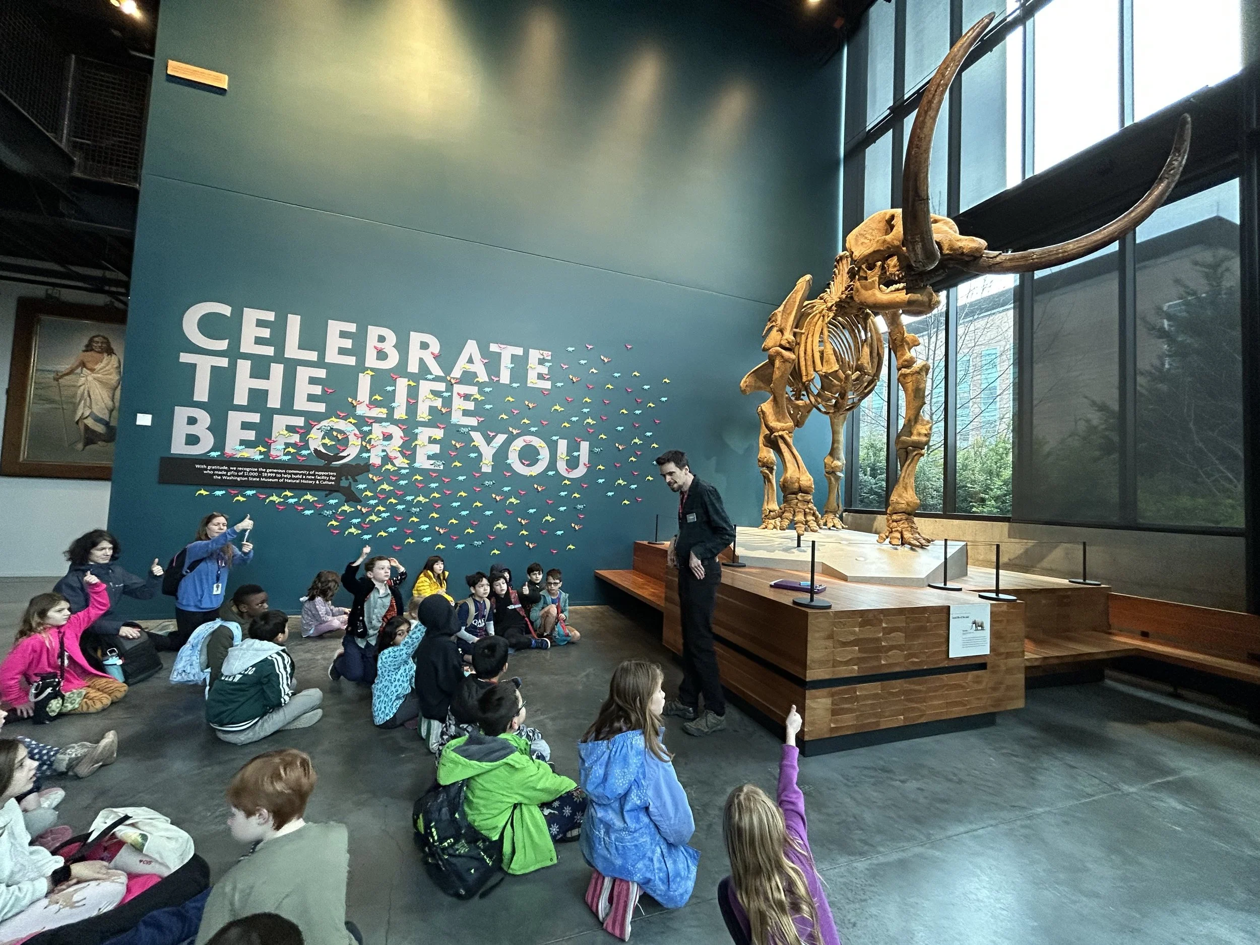 Children seated on the floor attending a presentation about a large dinosaur skeleton displayed behind them at a museum.