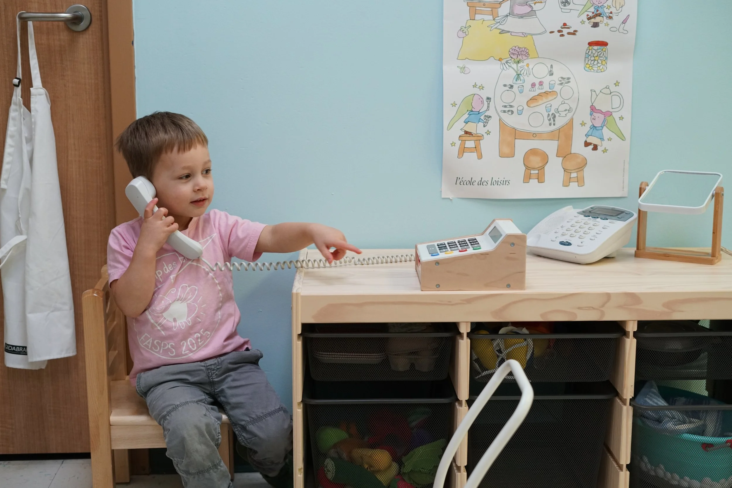 A young boy sitting at a wooden desk on a chair with a telephone headset, pointing at a computer keyboard, with a colorful poster on the wall behind him. The poster depicts a whimsical scene of children at a picnic and playing, with the text "l'école des loisirs".