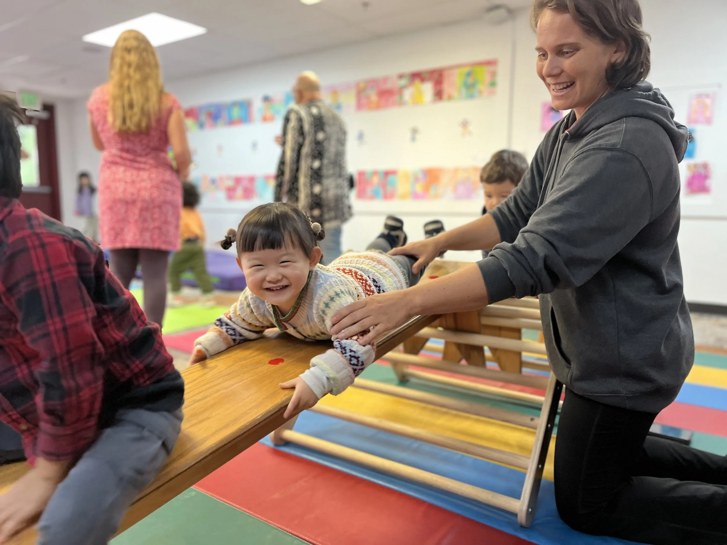 A young girl with pigtails smiling while lying on her stomach on a wooden table, being supported by a woman in a gray hoodie during a group activity in a colorful classroom.