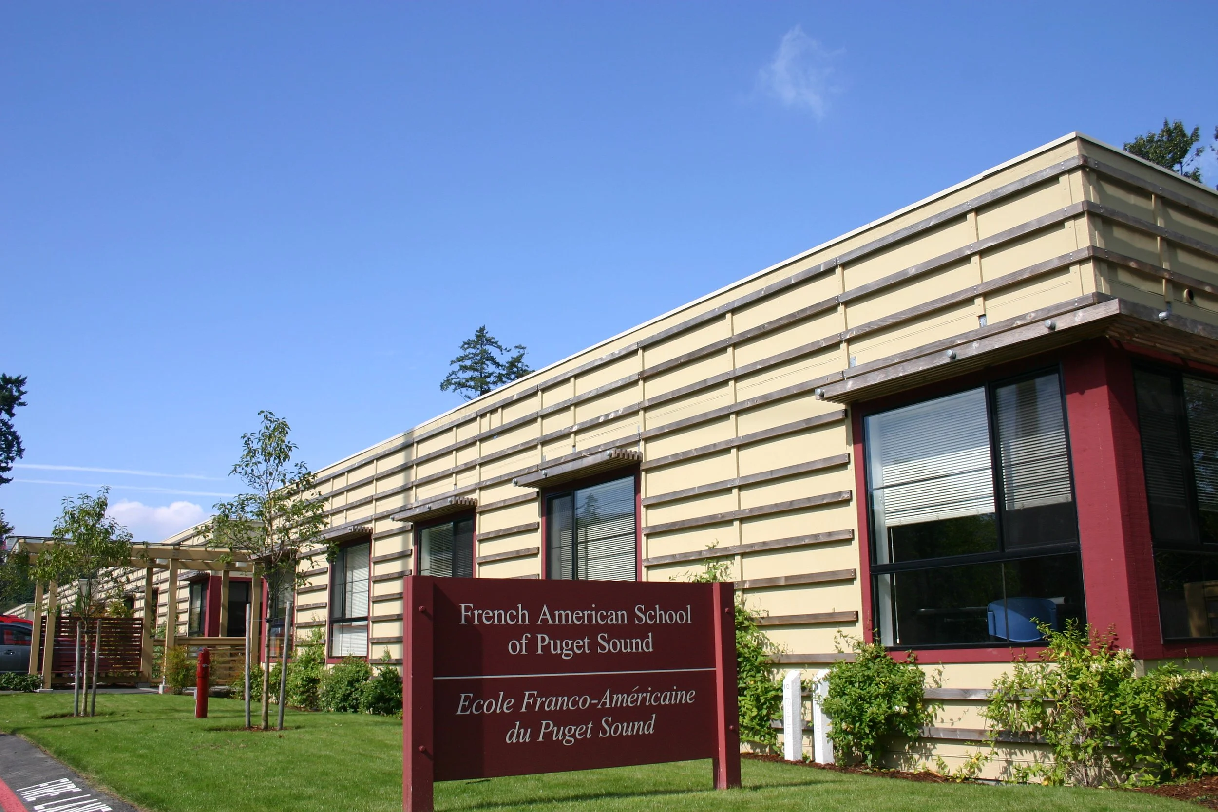 A building with a red sign that reads 'French American School of Puget Sound' and the French translation below. The building has beige siding with horizontal wooden slats, large windows, and a small landscaped area with trees and shrubs in front. The sky is clear with a few clouds.