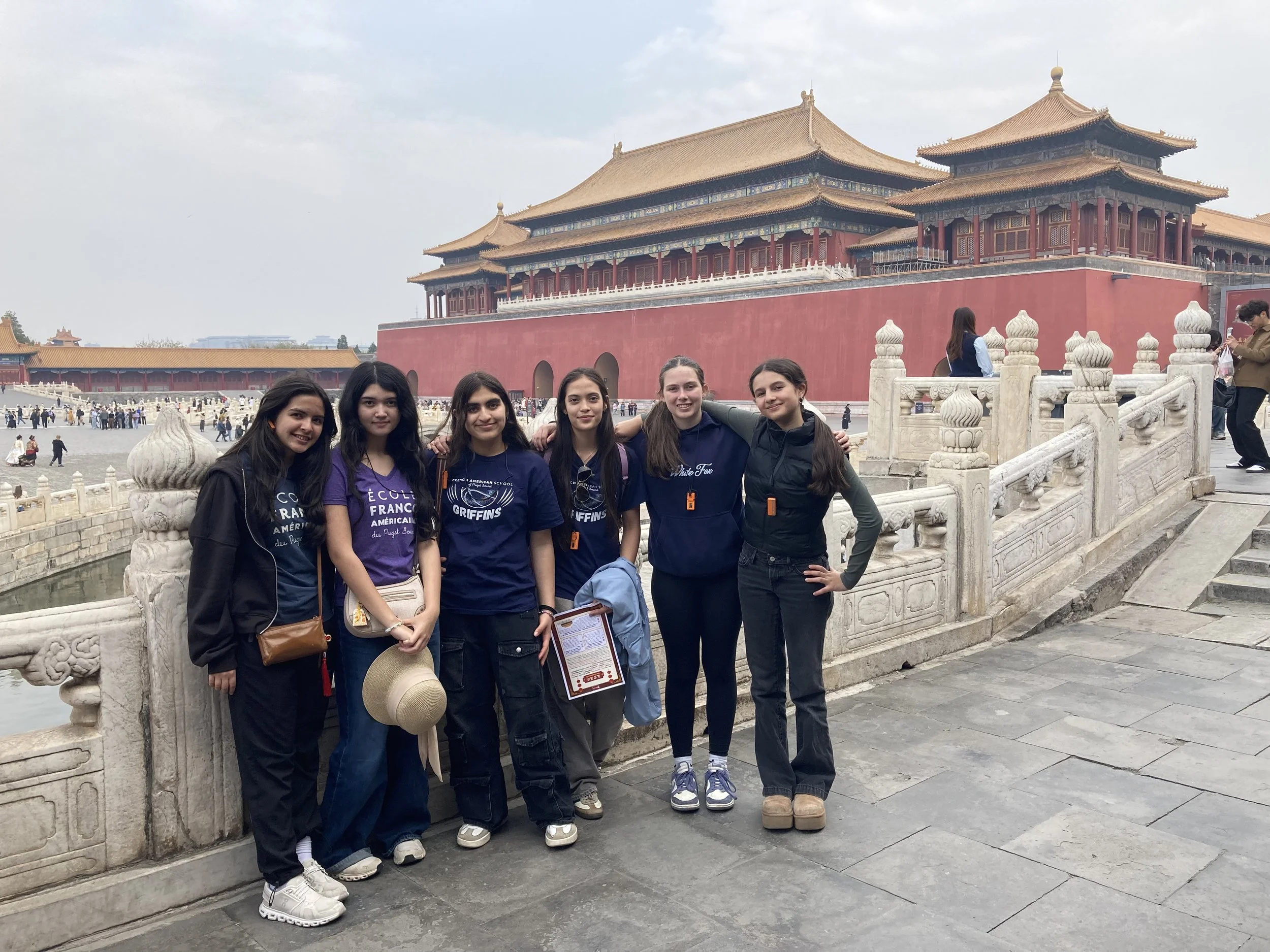 Group of six young women standing in front of a historic Chinese building with traditional architectural features such as ornate rooflines, red walls, and stone railings. They are smiling and appear to be tourists enjoying a visit to a cultural site.