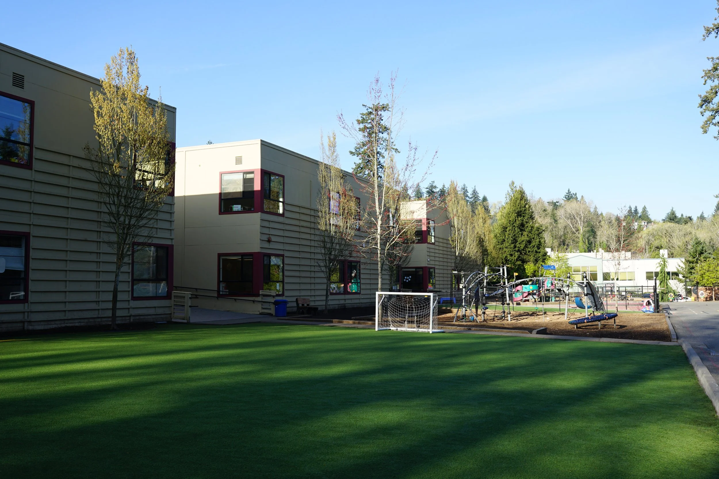 A school playground with a large, neatly maintained green grass field, surrounded by trees, metal playground equipment, and a soccer goal. In the background are modern school buildings with large windows and a partly cloudy sky.