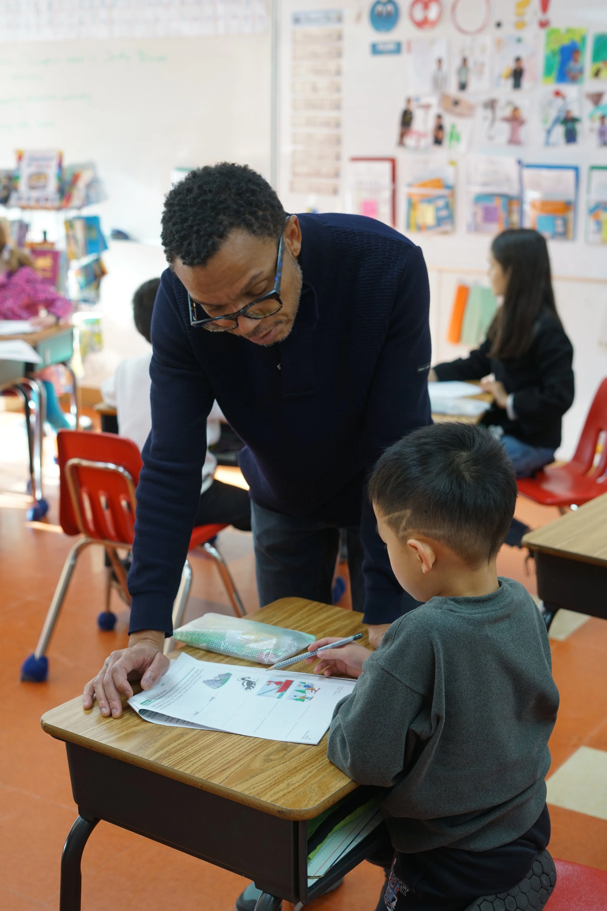 An adult male teacher leaning over a student's desk, assisting a young student with a worksheet in a classroom. Other students are seated at desks in the background.