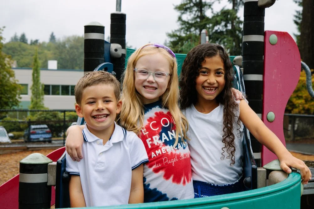 Three children smiling and posing together on a playground structure, outdoors in a park with trees and a building in the background.