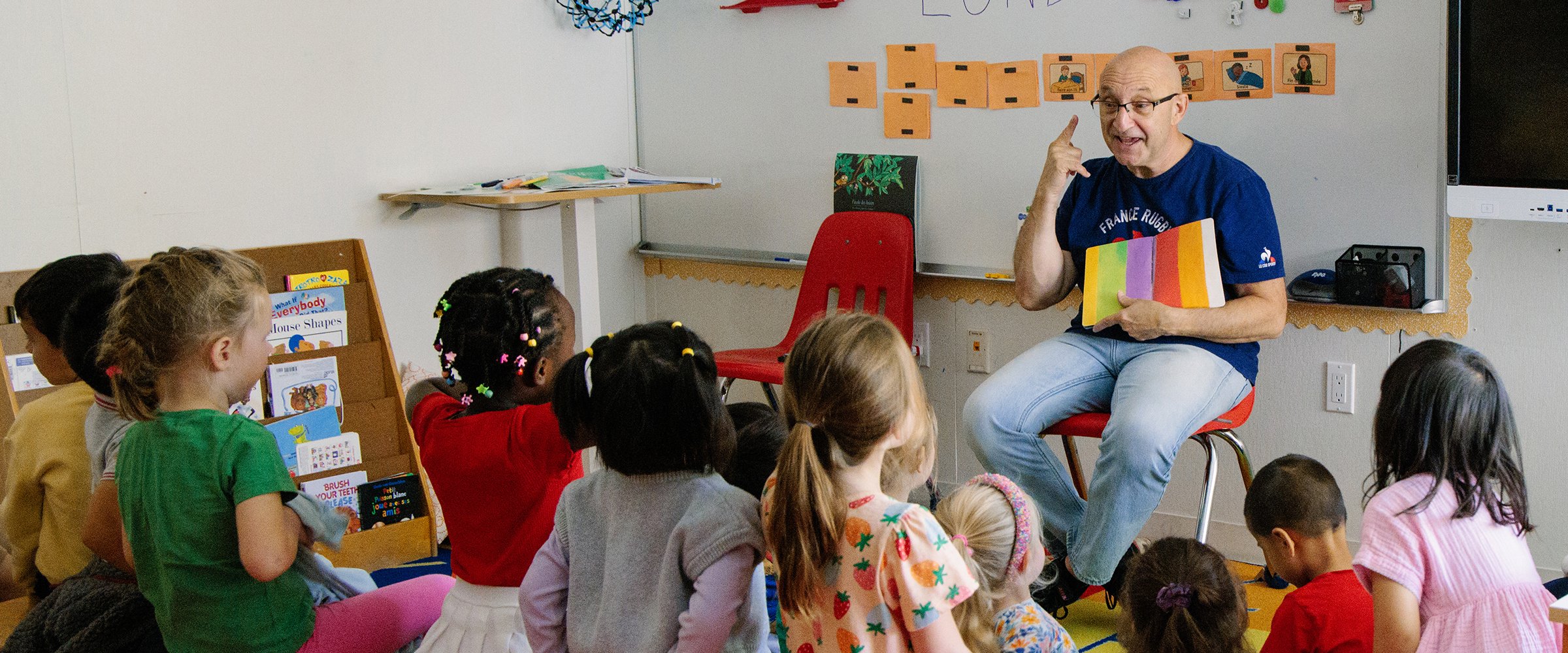 A classroom scene with a teacher reading to a diverse group of children seated on the floor. The teacher is sitting on a red chair, holding a colorful book and gesturing as he speaks. The children are attentively listening, some with their hands on their laps. There are educational materials and posters on the walls and shelves, including a display with books and a whiteboard behind the teacher.