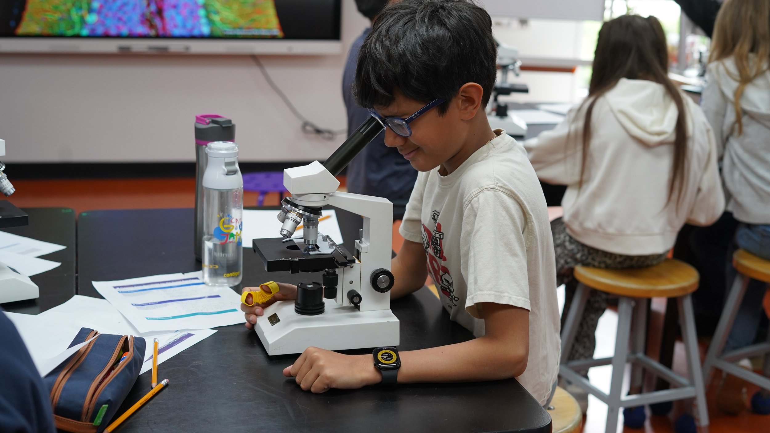 A boy wearing glasses looking through a microscope in a classroom setting.