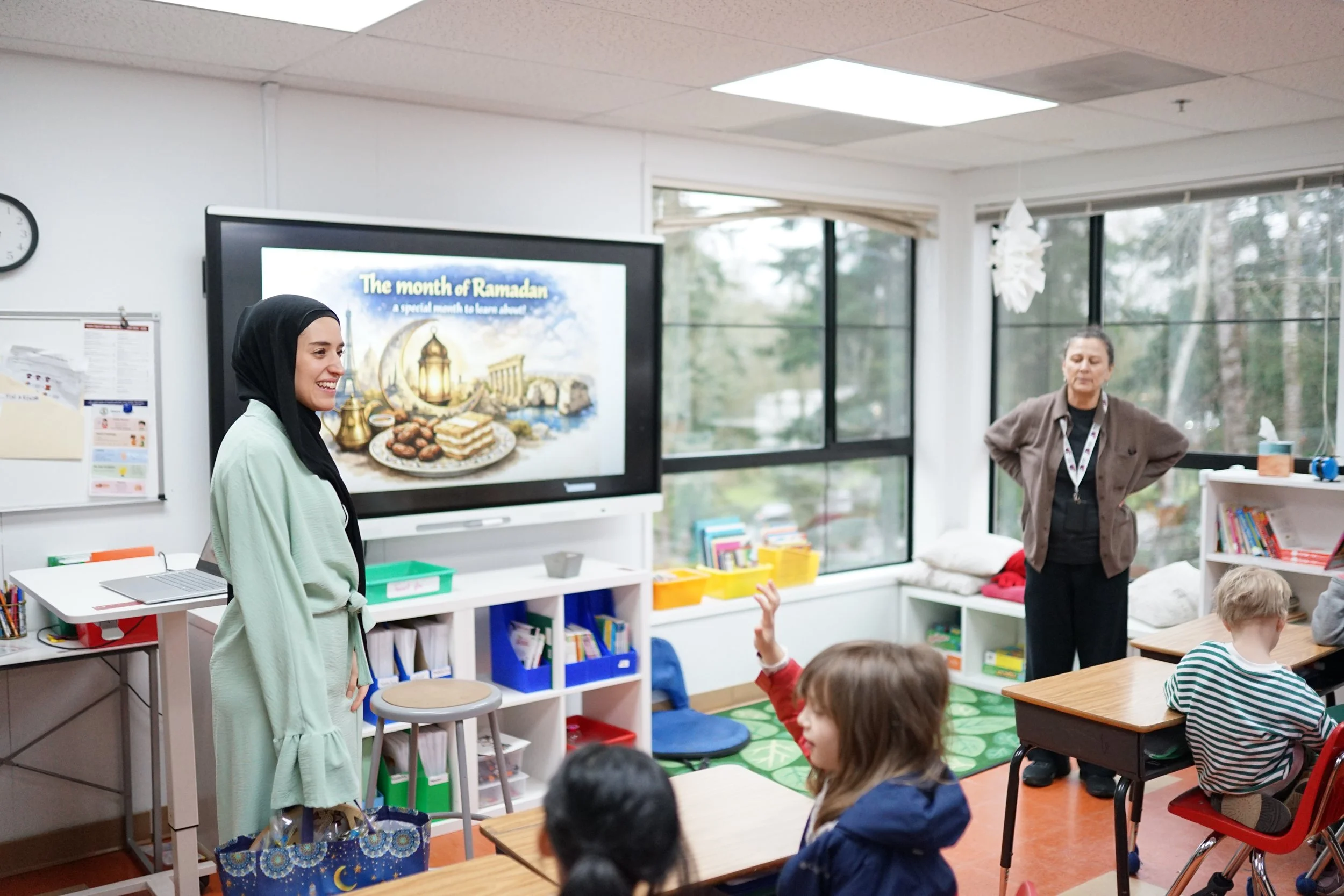 A classroom with children sitting at desks, a woman standing with her hands on her hips, and a woman smiling near a large screen displaying a slide about Ramadan. The classroom has large windows, bookshelves, and educational posters.