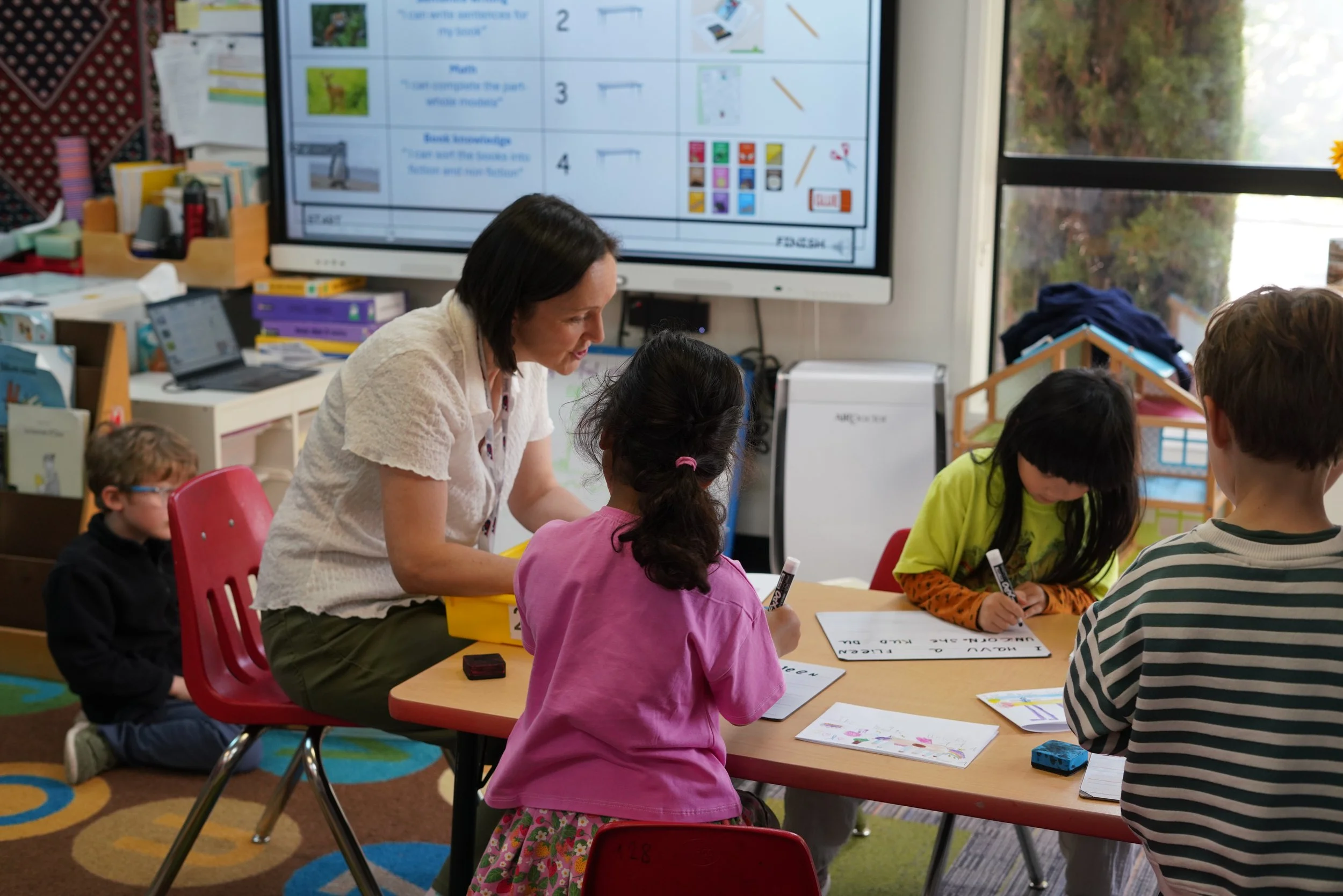 A teacher assisting young students with educational activities in a classroom. There is a large digital screen in the background displaying educational content, and the students are seated at a table writing on paper or whiteboards.