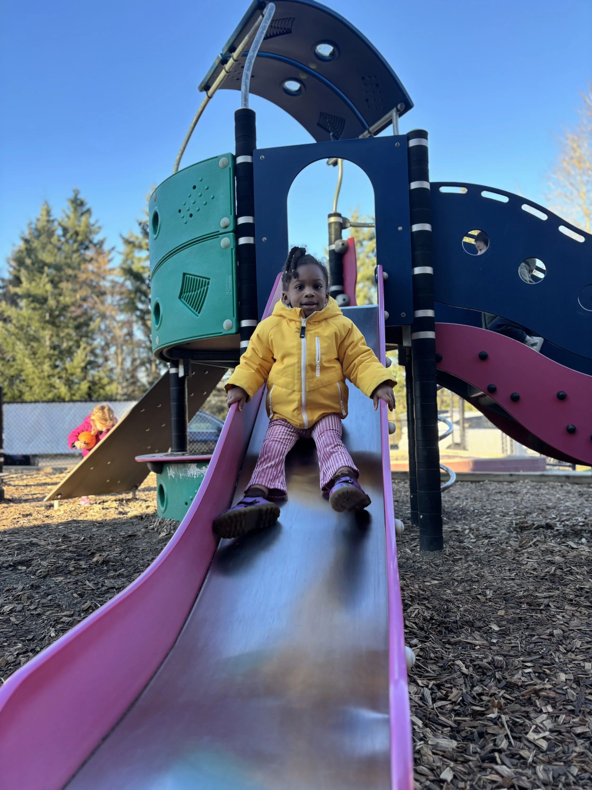 A young girl wearing a yellow jacket and pink striped pants sliding down a playground slide at a park.