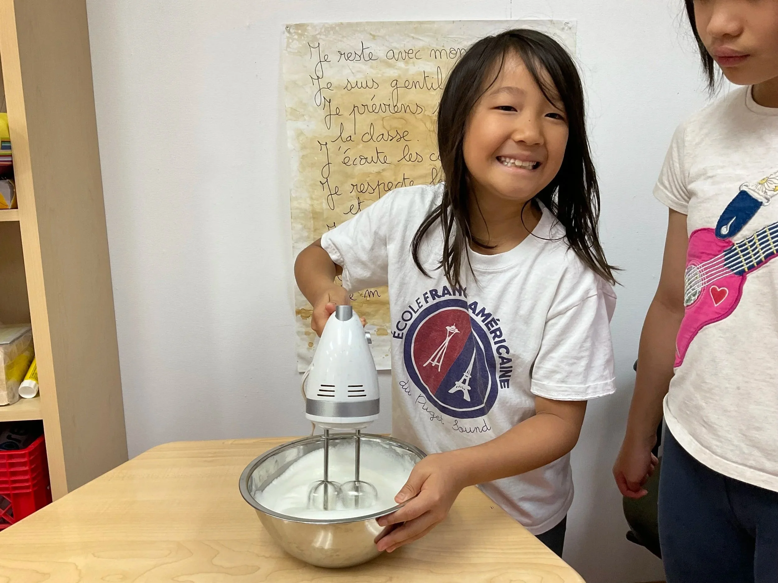 A girl is using a cotton candy machine to make cotton candy, wearing a white T-shirt with a logo, while another girl stands nearby. Behind them is a poster on the white wall with French text.