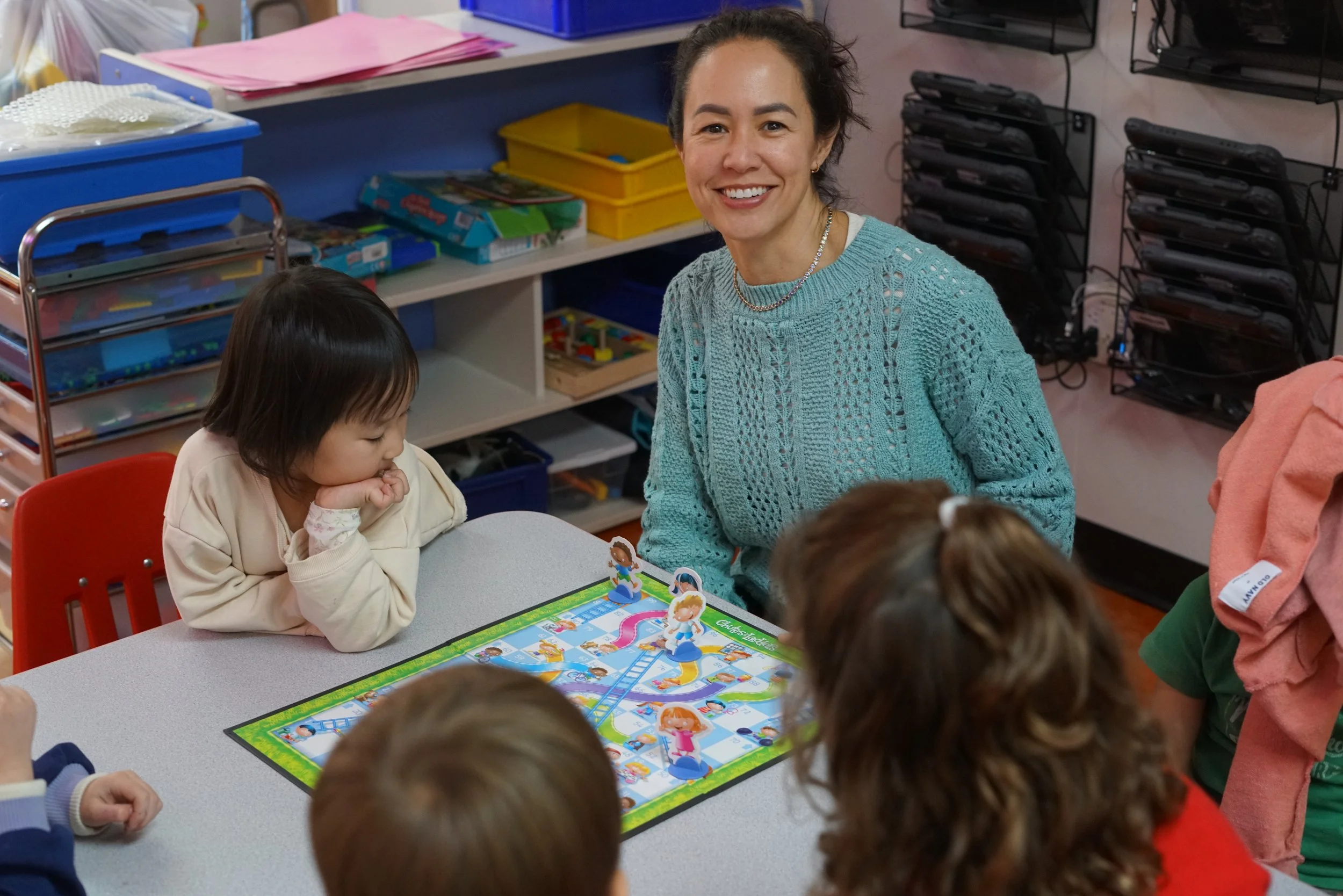 A woman with short dark hair smiling at children around a table playing a board game in a classroom.