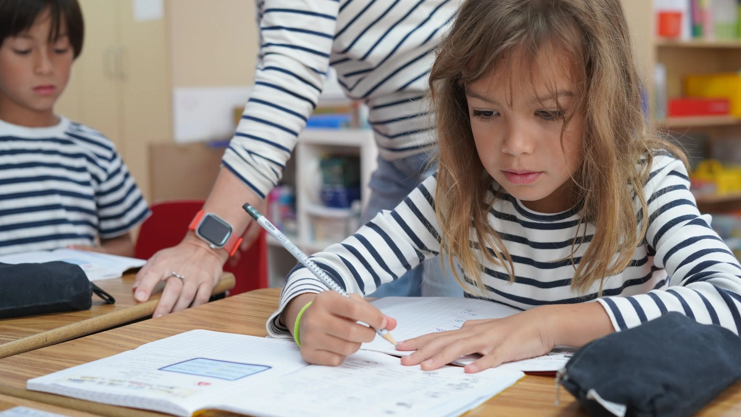 A young girl with long, curly hair and a striped shirt is sitting at a desk, writing in a notebook.